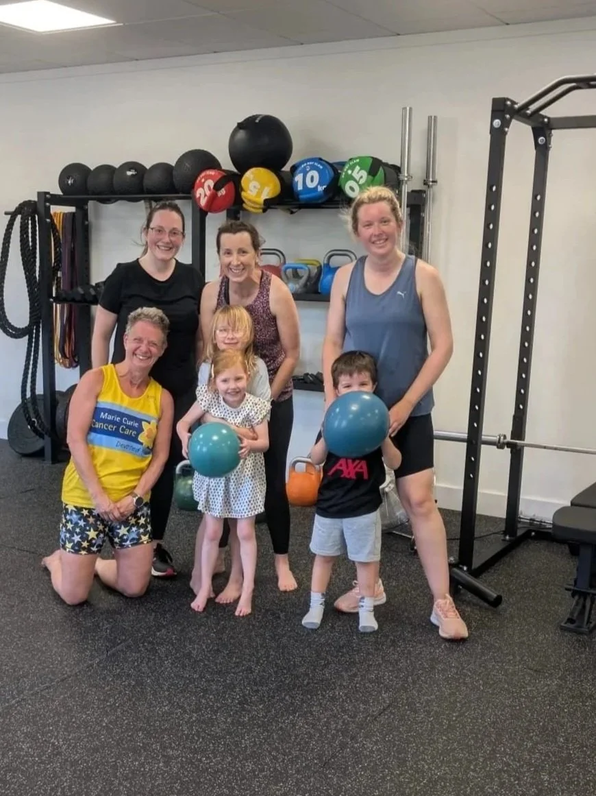 Group of seven people, including three children, posing in a gym with fitness equipment and black rubber flooring.