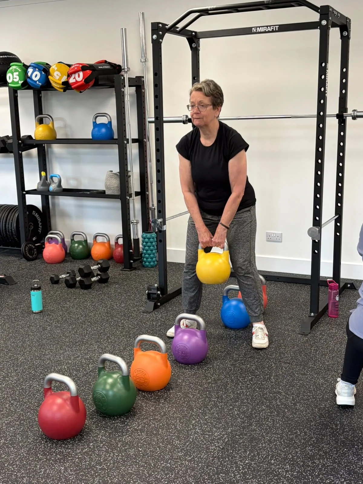 A woman lifting a yellow kettlebell in a gym, surrounded by colorful kettlebells and exercise equipment.