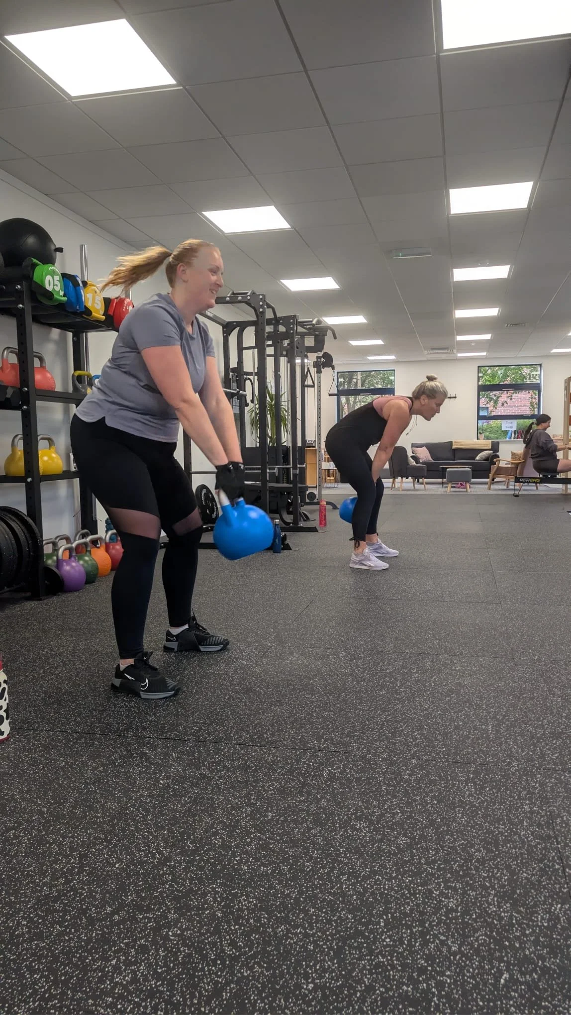 Three women exercising in a gym, lifting kettlebells and bending forward at the waist, with gym equipment and exercise balls in the background.