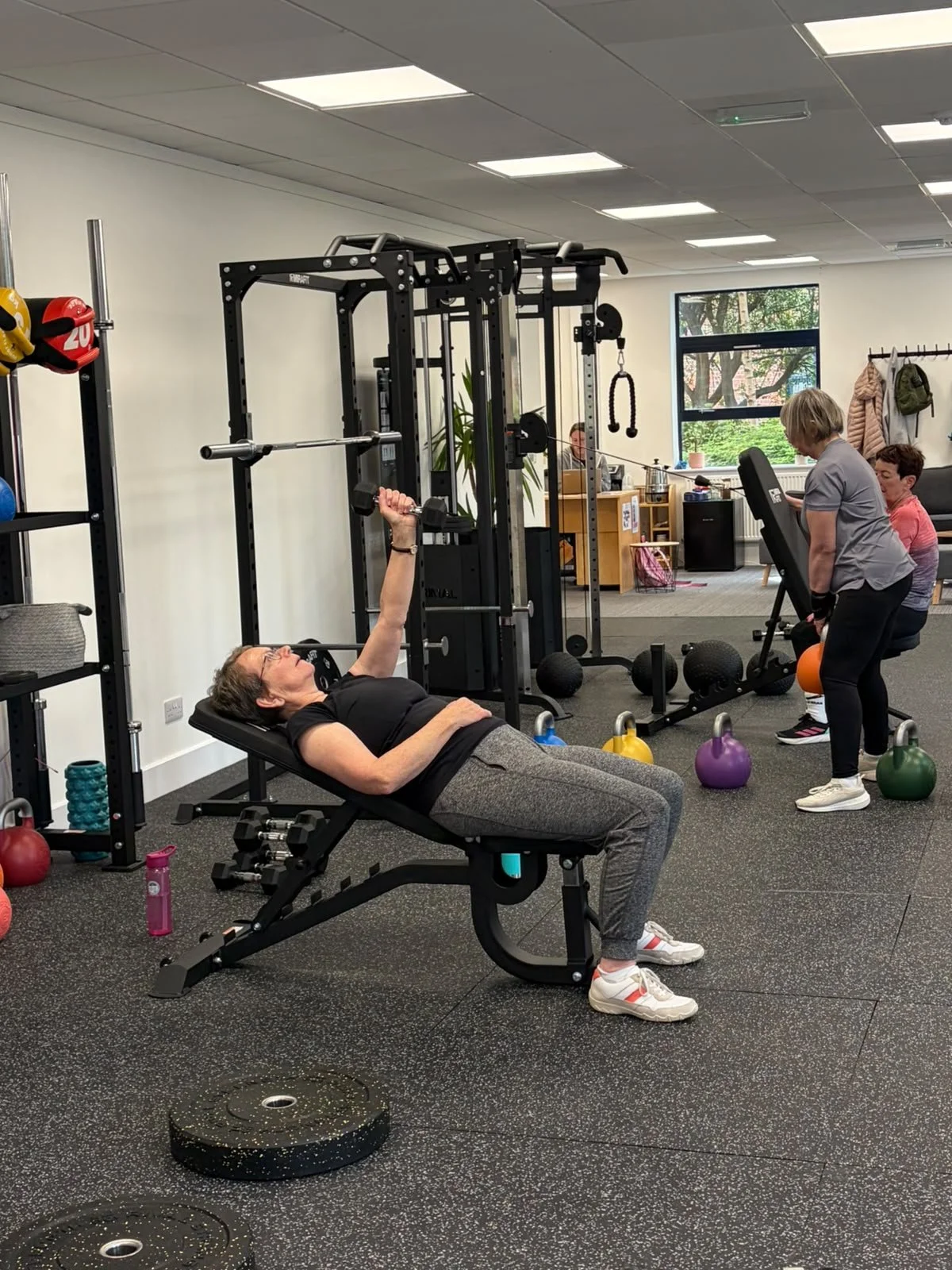 An woman lying on a workout bench in a gym, lifting a dumbbell, while other women have a kettlebell workout nearby. The gym has various weights and exercise equipment.