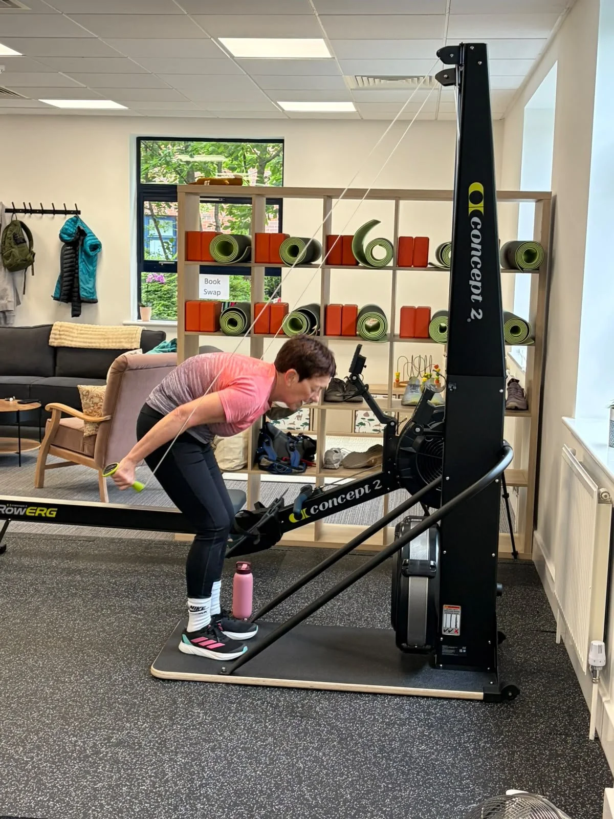 A person exercising on a rowing machine in a fitness room, with a water bottle nearby, a couch, and a shelf with foam rollers and other gear in the background.