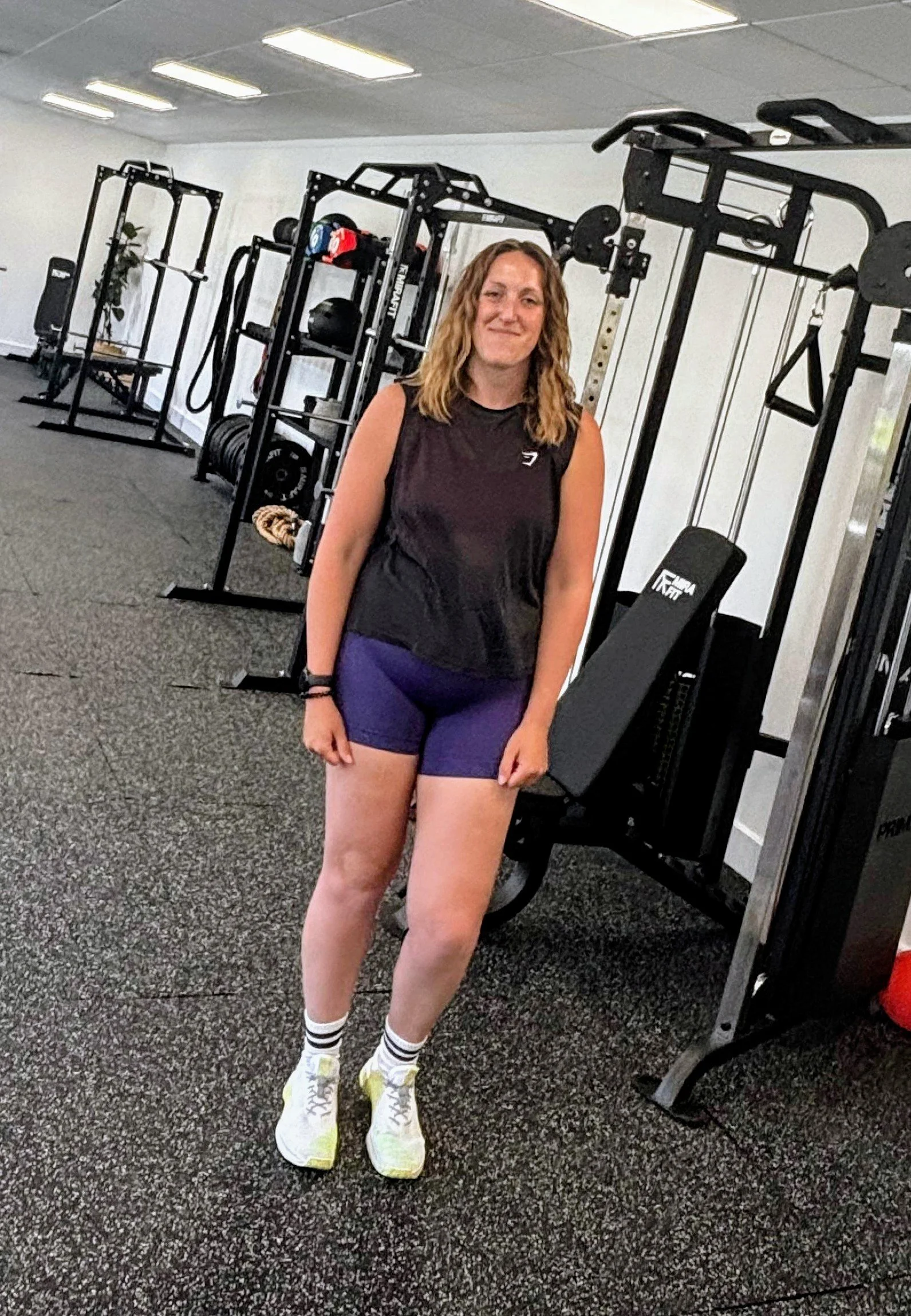 A woman with curly brown hair in workout attire standing in a gym with workout equipment and a mirror in the background.