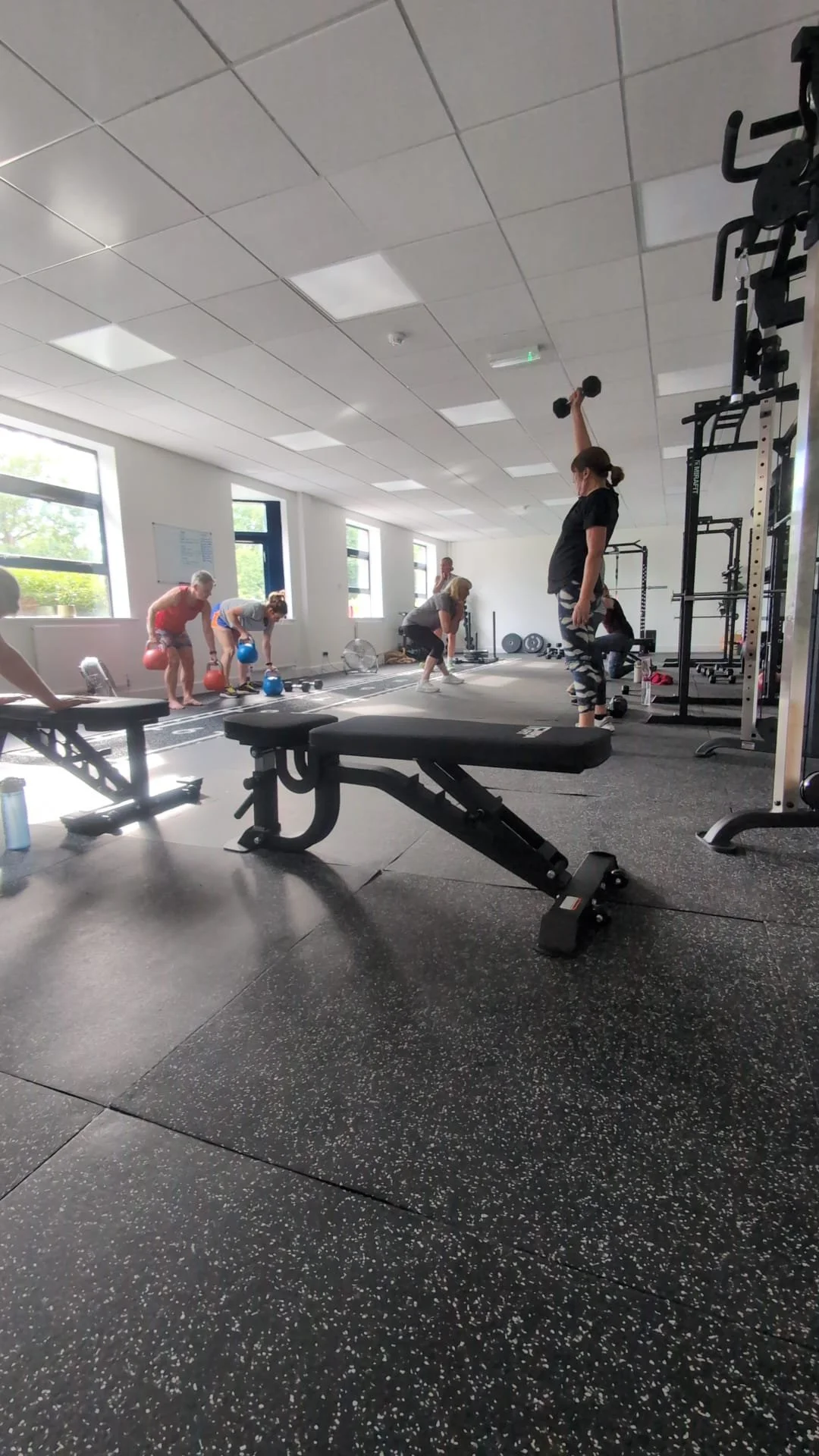 People working out in a gym, performing exercises with kettlebells and dumbbells, with a woman lifting a dumbbell overhead.