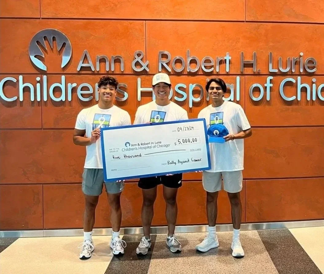 Three young men standing in front of a wall with 'Ann & Robert H. Lurie Children's Hospital of Chicago' sign, holding a large check for $5,000 made out to the hospital, with the memo 'Rally Against Cancer'.