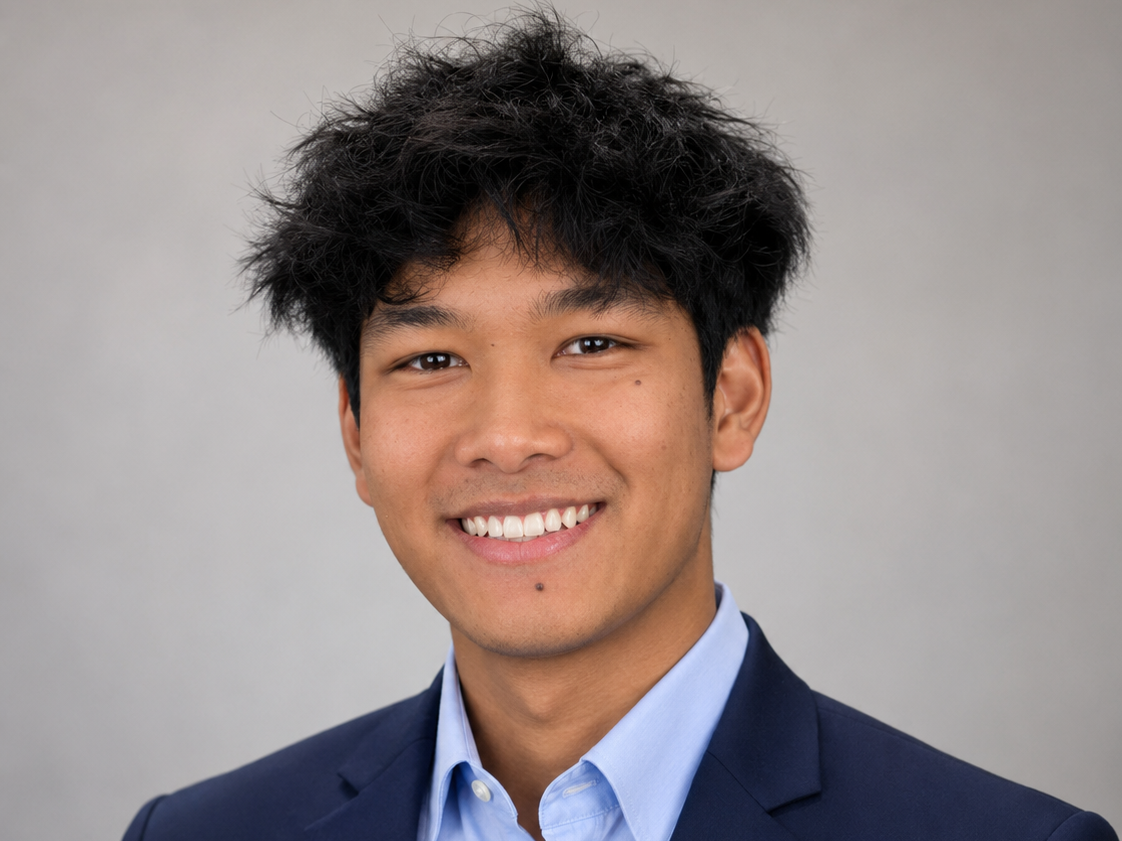 Headshot of a young man with black hair, wearing a navy blazer and light blue dress shirt, smiling against a plain grey background.