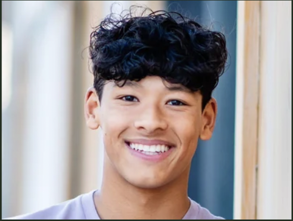 Young man with curly hair smiling in an outdoor setting.