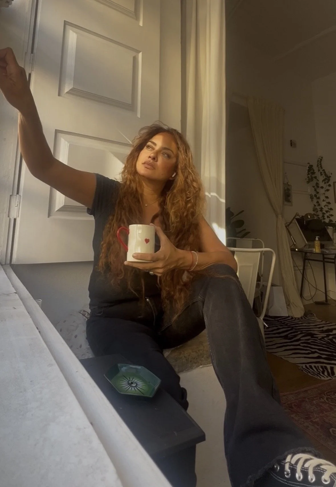 A woman with long, curly hair sitting on the floor in a room, holding a white mug with a red heart, looking off to the side. The room has a white door, beige curtains, a zebra-striped rug, and a small table with a lamp and decorative plant.