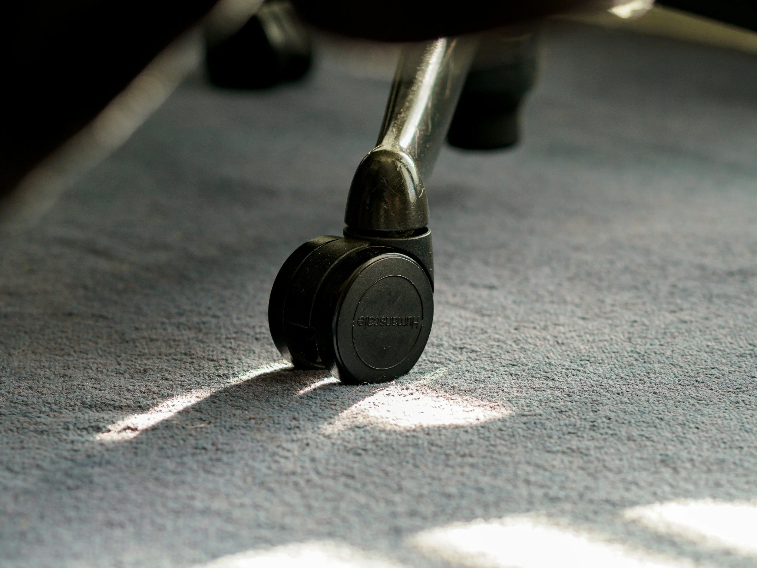 Close-up of a caster wheel attached to furniture, with sunlight casting shadows on a carpeted floor.