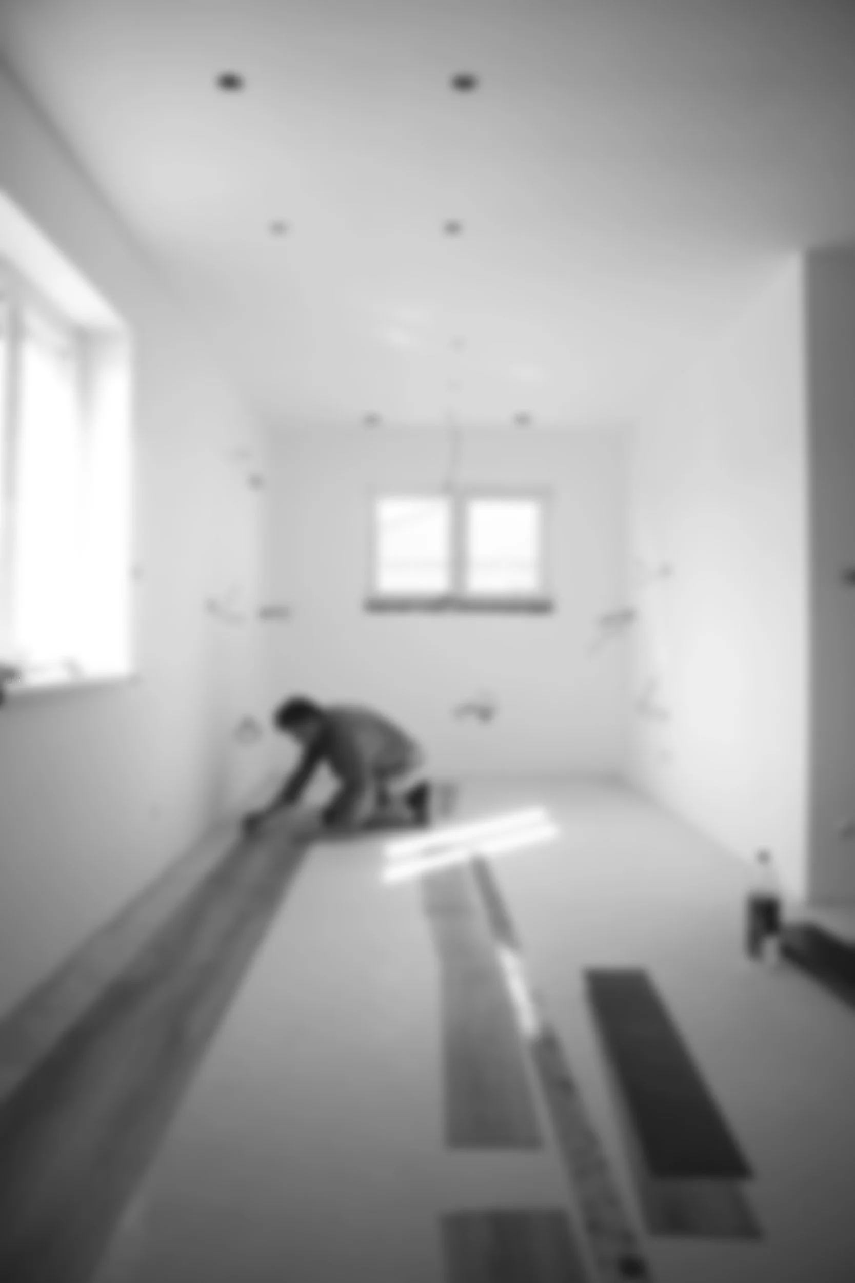 Black and white photo of a person using a spirit level to check the levelness of a wooden plank in a room under construction or renovation.
