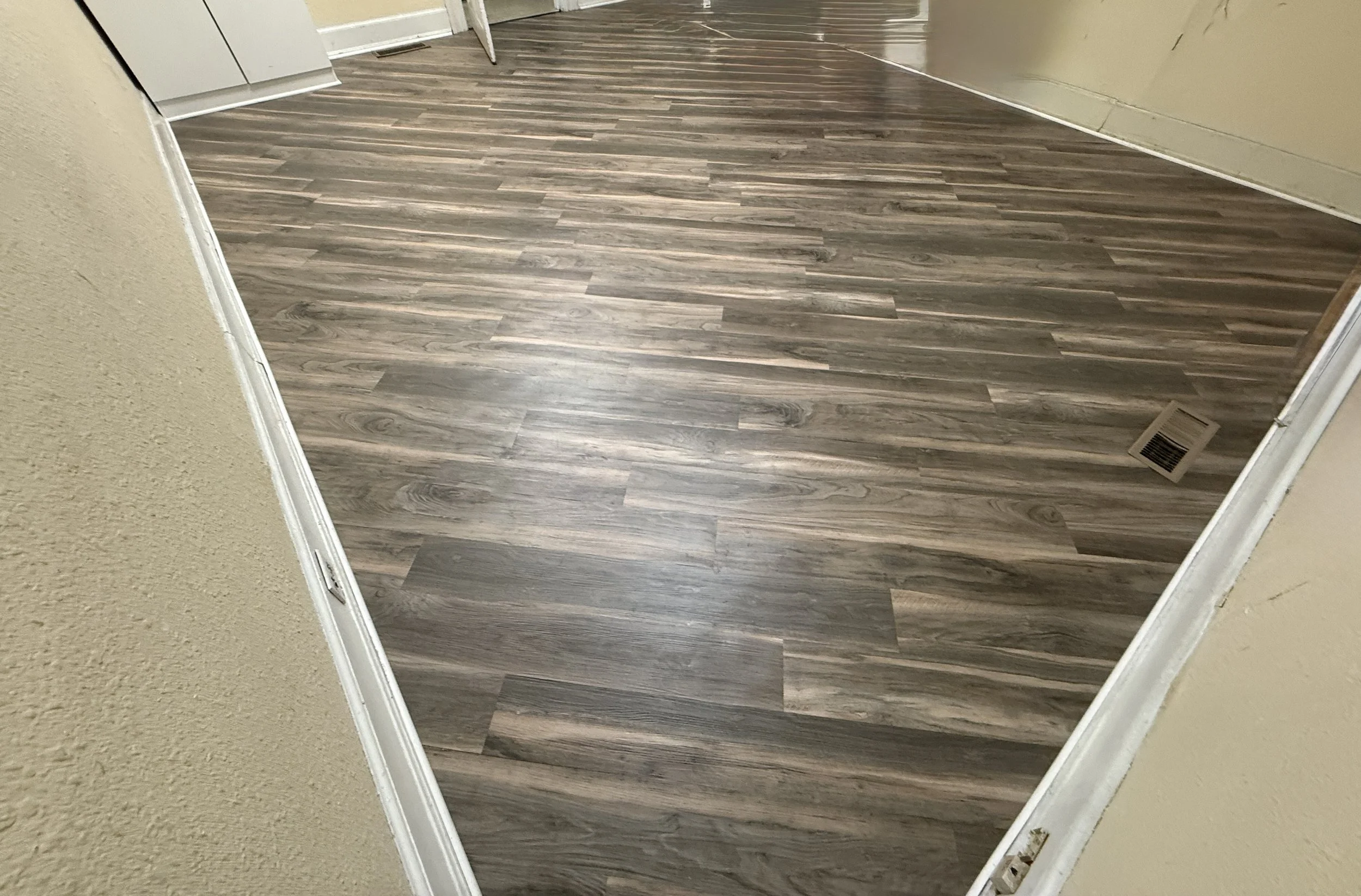 Interior view of a room showing wood laminate flooring, beige wall to the right, and a partial view of a kitchen with white cabinets on the left.