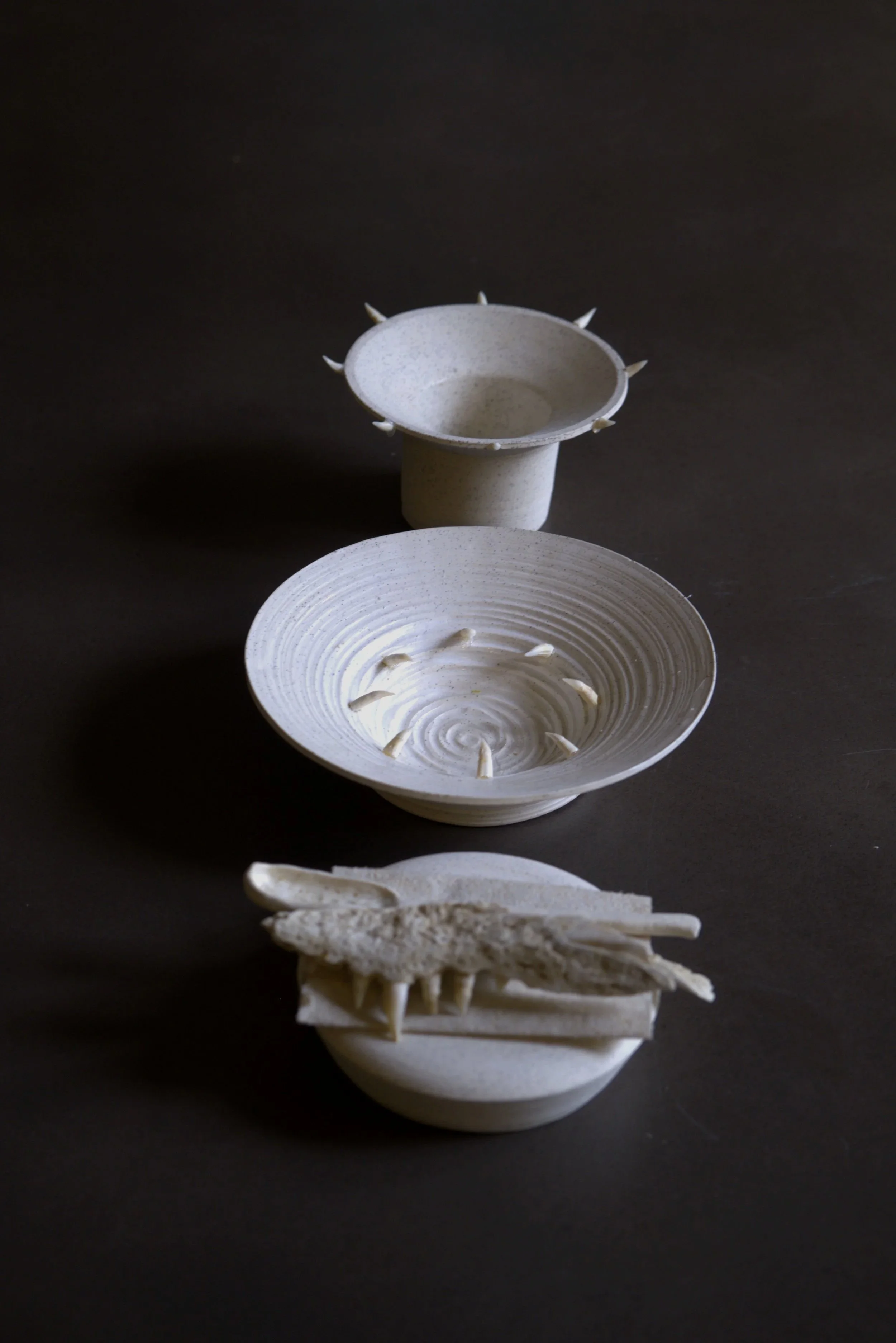 A ceramic vessel with small spike-like protrusions along the rim, placed on a black surface with a second open bowl and a white decorative object resembling a bird in front.