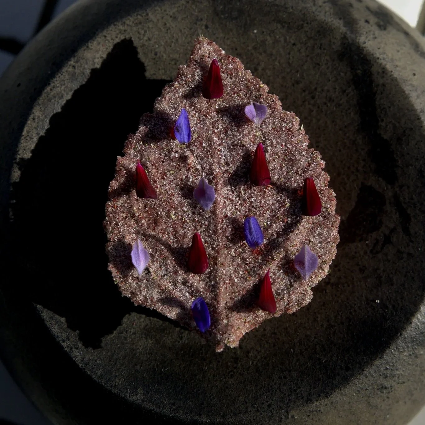 A pyramid-shaped cake decorated with red and purple flower petals, topped with sugar crystals.
