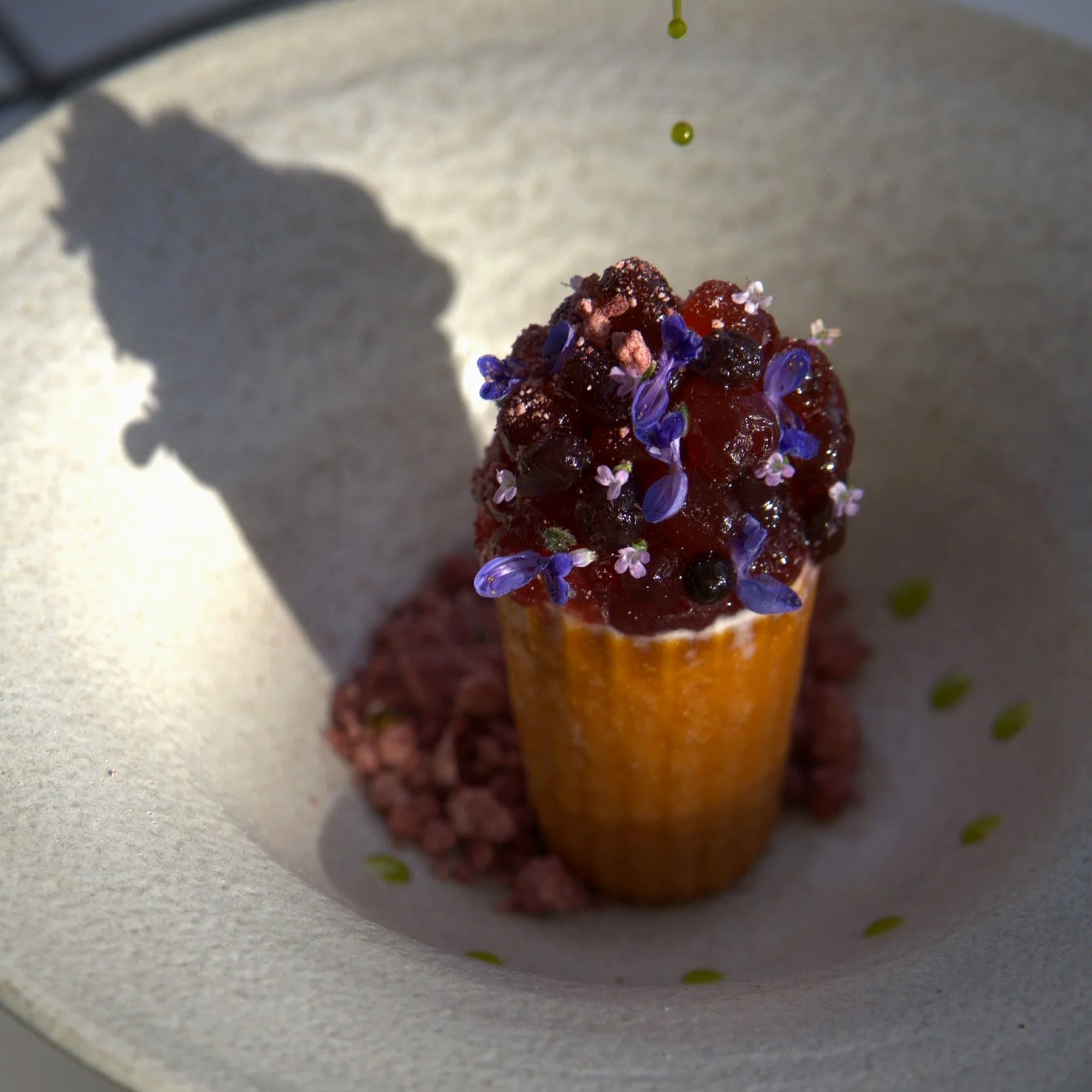 Close-up of a dessert ice cream cone topped with berry compote, edible flowers, and sprinkled with pink crumbs, placed on a white plate with green dot pattern.