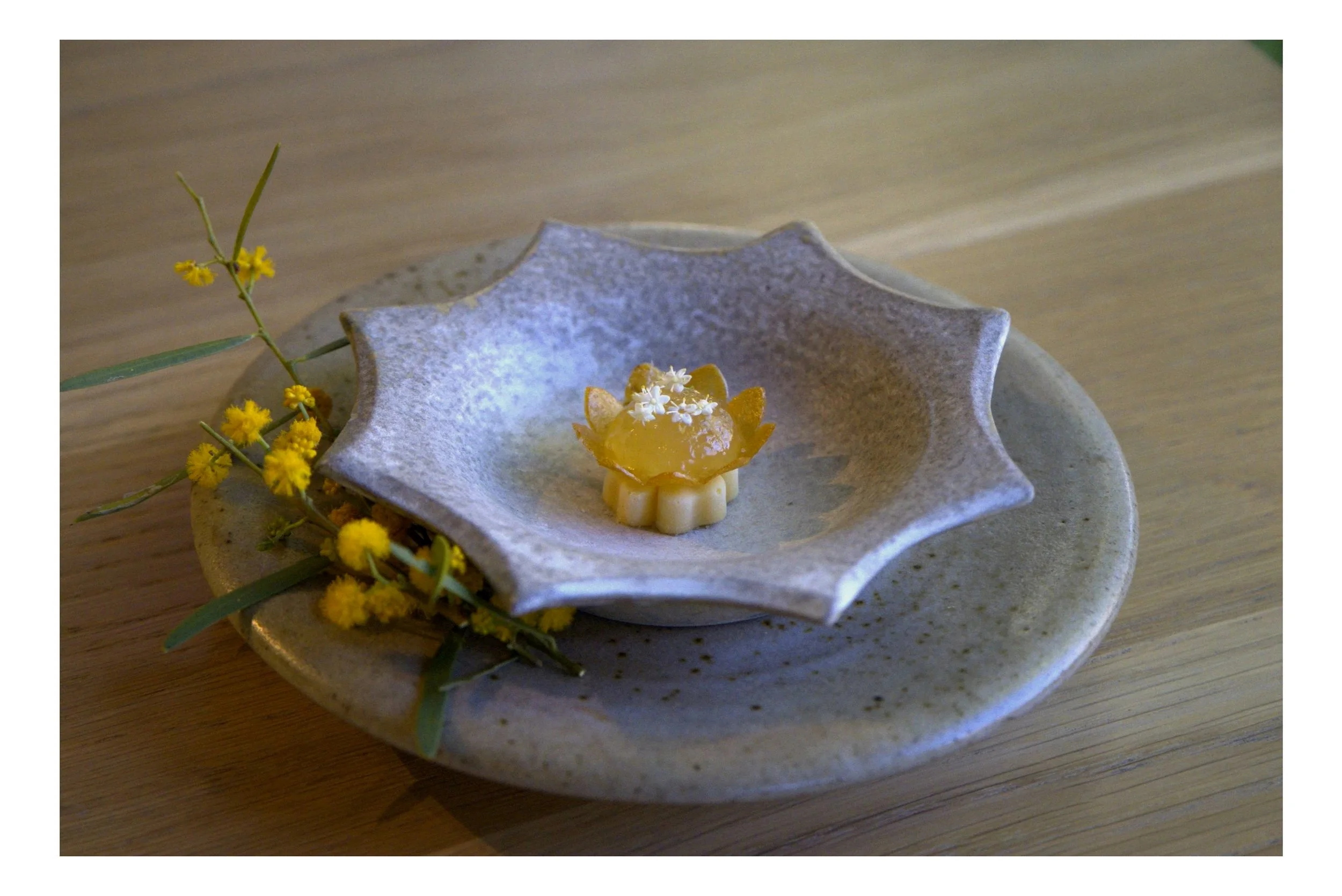A ceramic dish with a flower-shaped dessert topped with white flowers, placed on a larger ceramic plate with yellow flowers and green leaves next to it, on a wooden table.