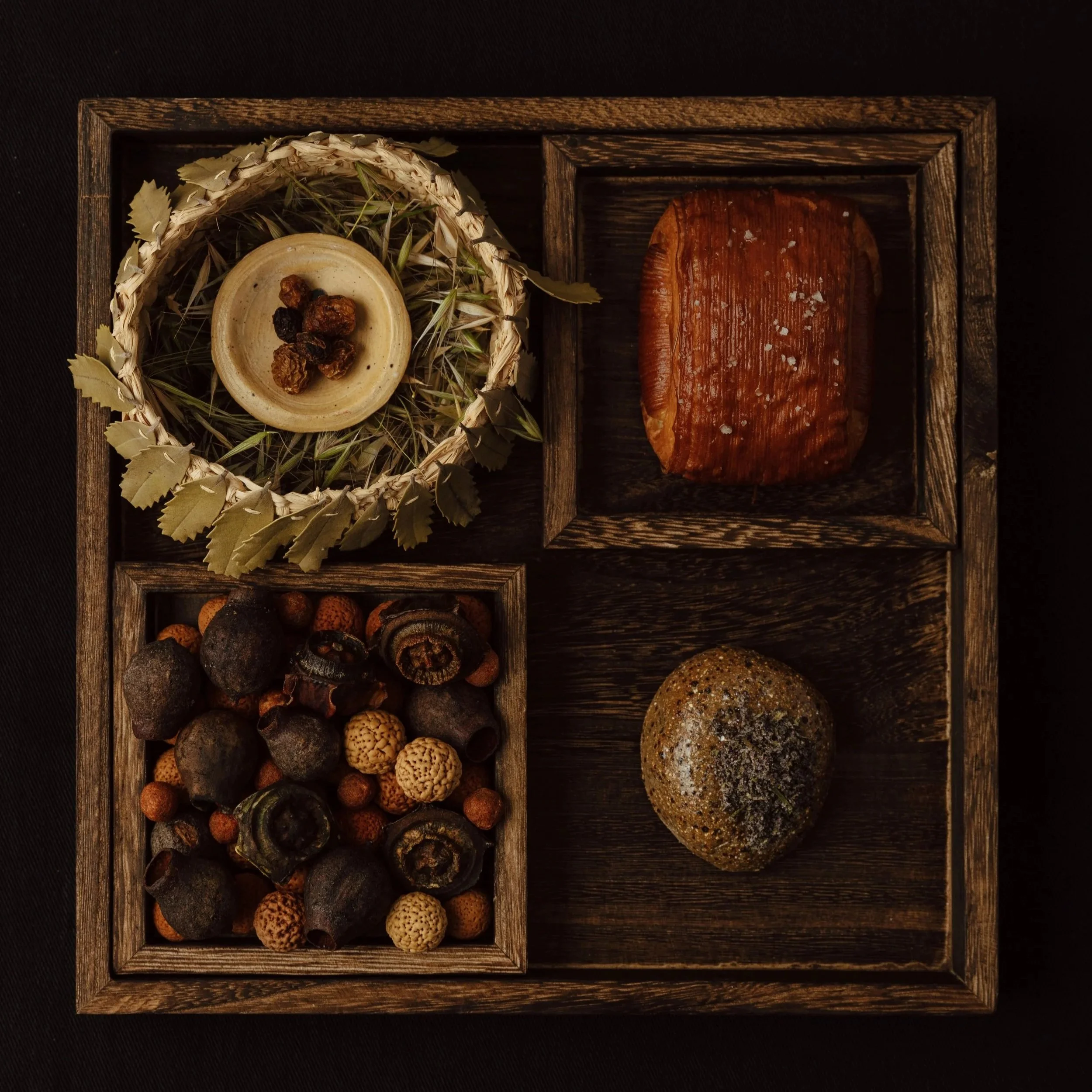 A wooden tray with separate compartments holding dried herbs, small dried fruits, dark dried mushrooms, and various round shapes, possibly nuts or seeds, on a dark surface.