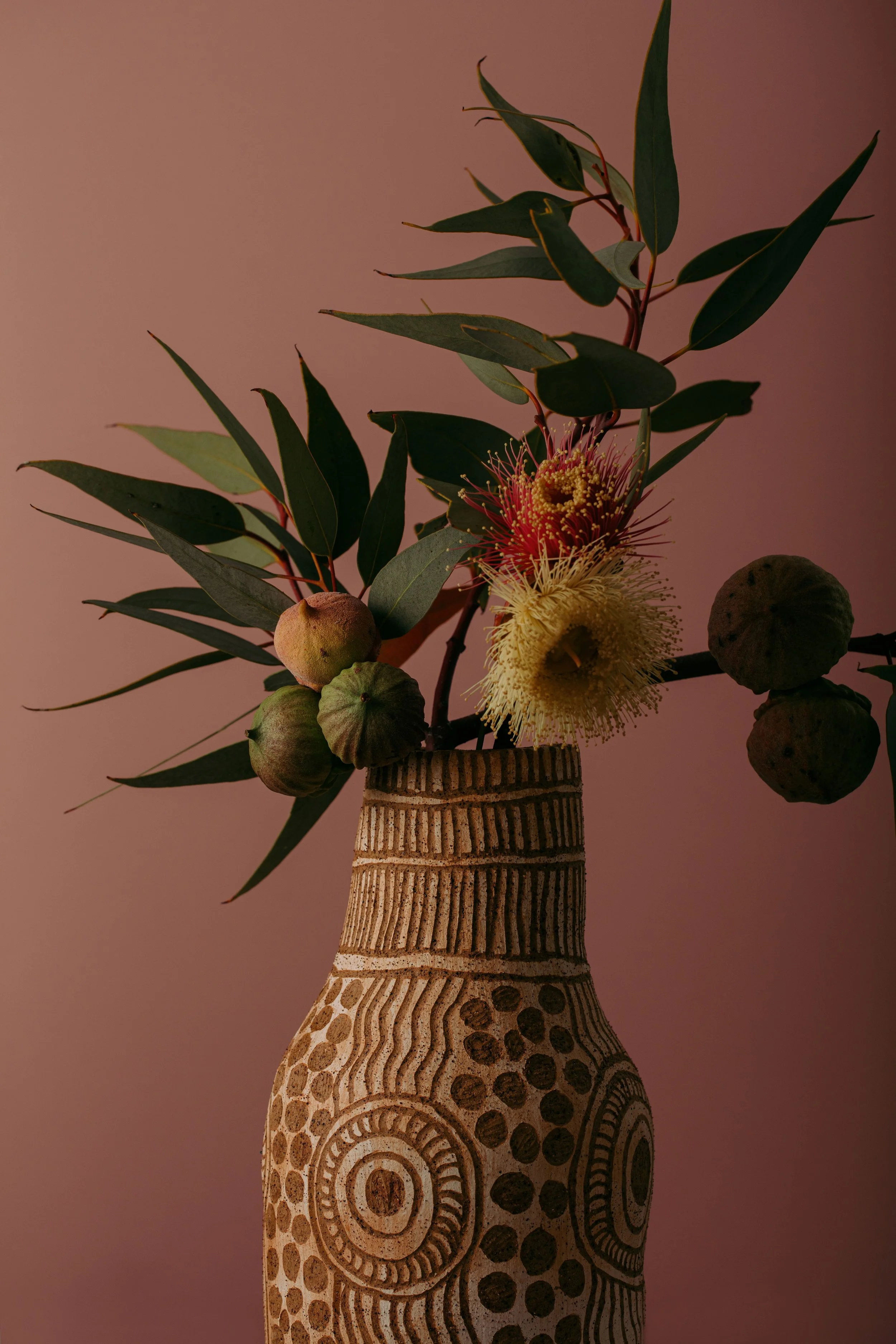 A decorative vase with a tribal pattern holding a green leafy branch with pink and yellow flowers, set against a plain brown background.