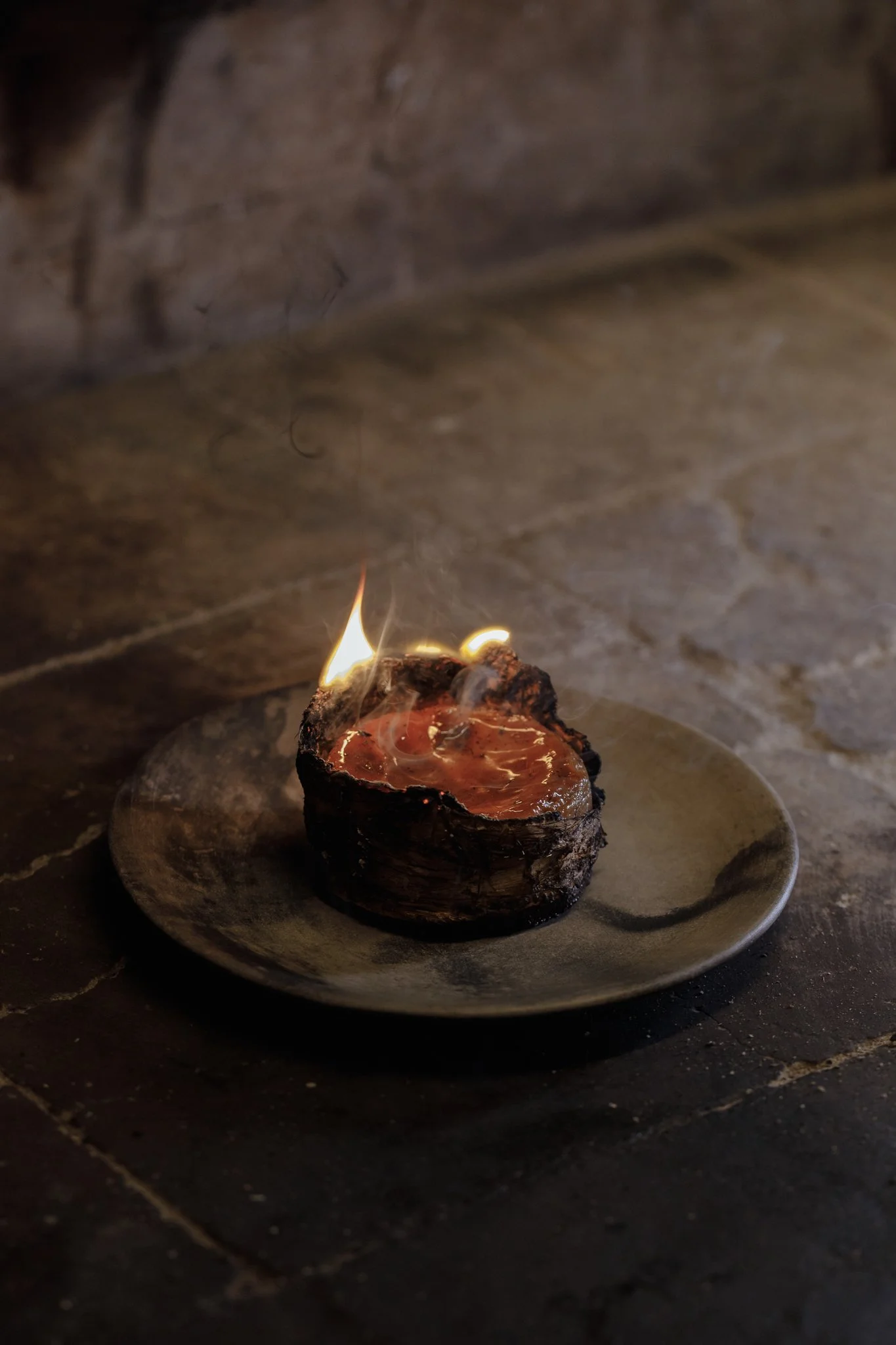 A small candle with a flame burning inside a hollowed-out piece of wood, placed on a round ceramic plate on a dark surface.