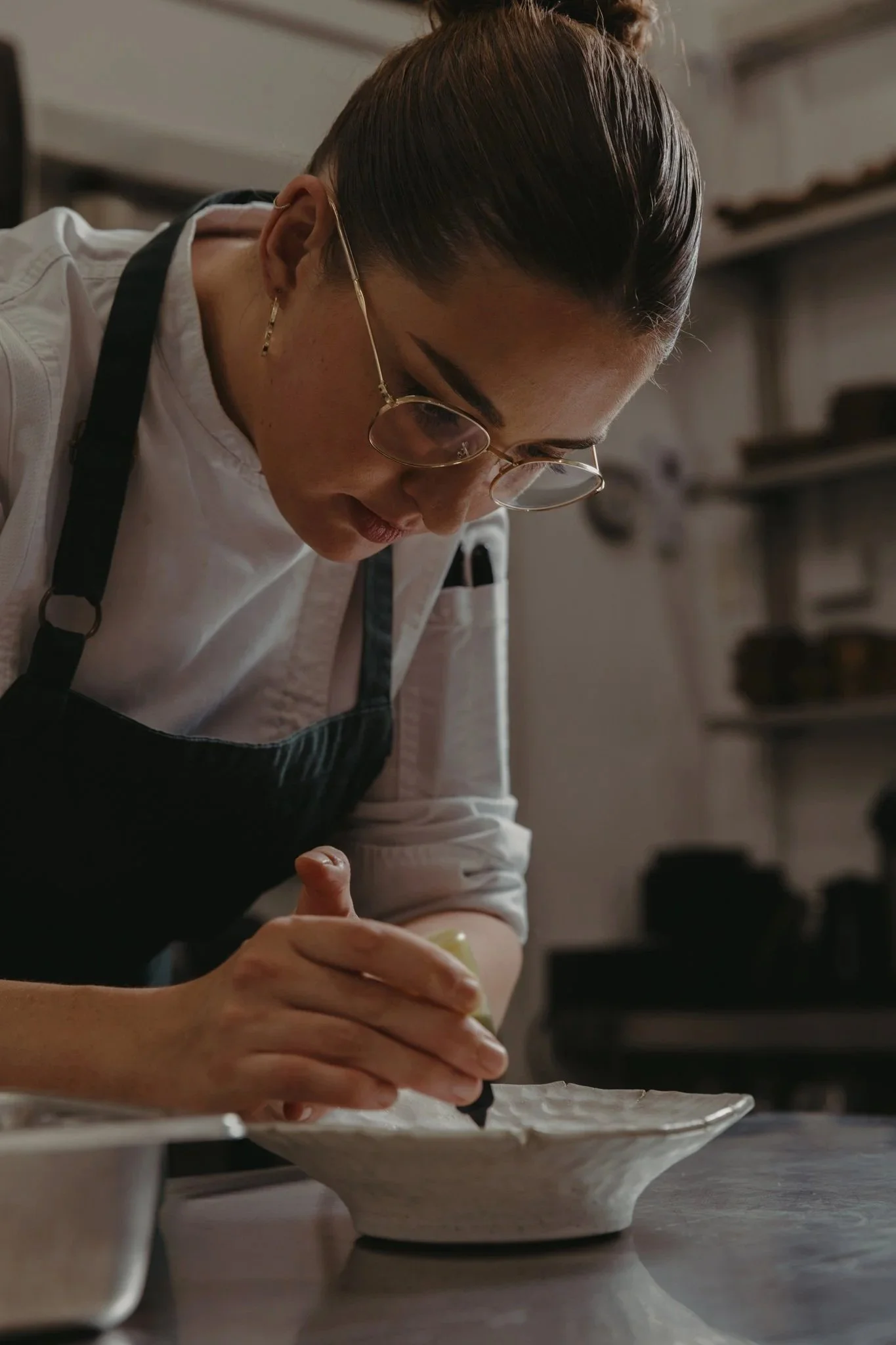 A woman with dark hair, glasses, and a white chef's coat is preparing food in a kitchen, holding a utensil over a plate.