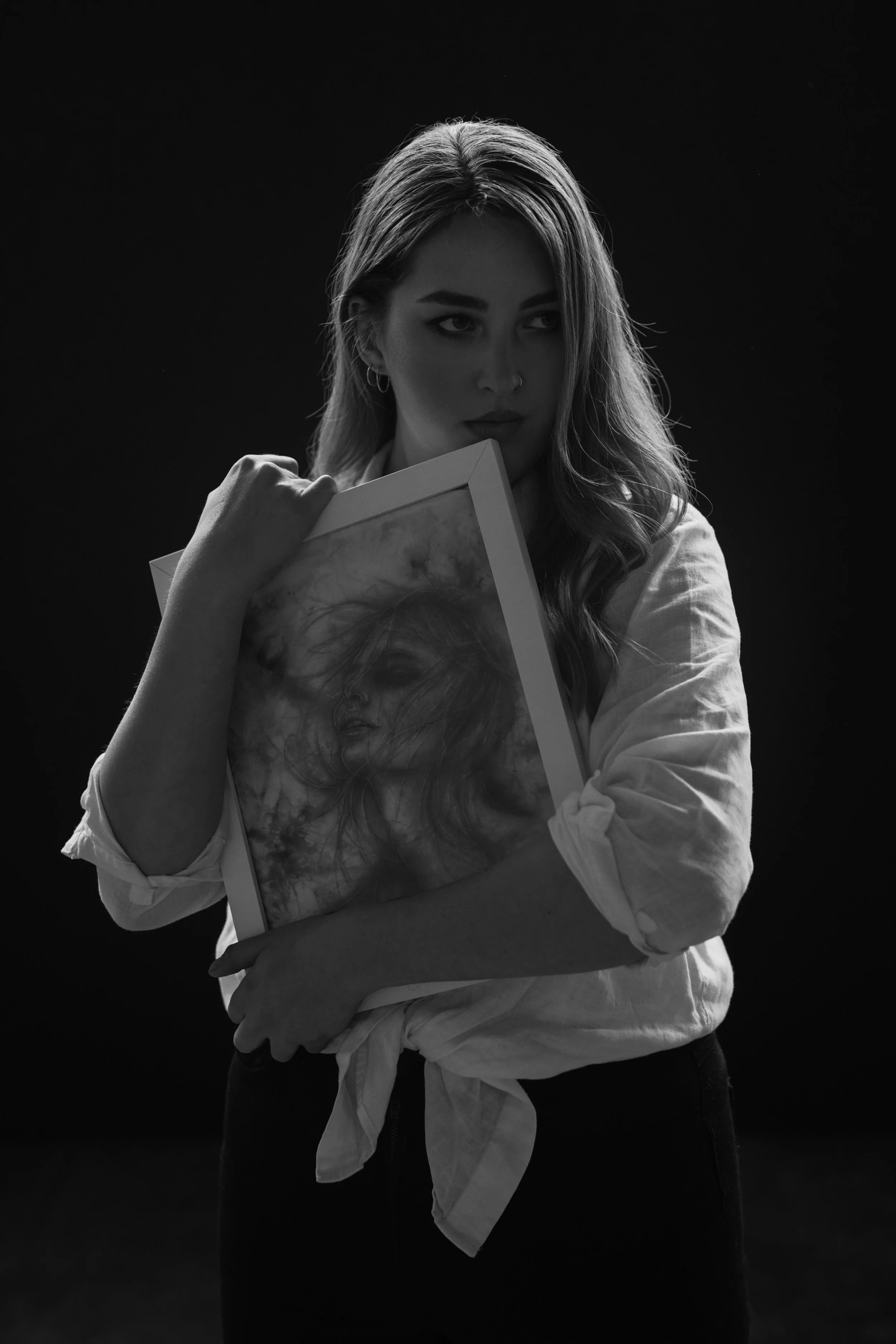 A woman with long wavy hair holding a framed artwork, lookings away thoughtfully, with a dark background.