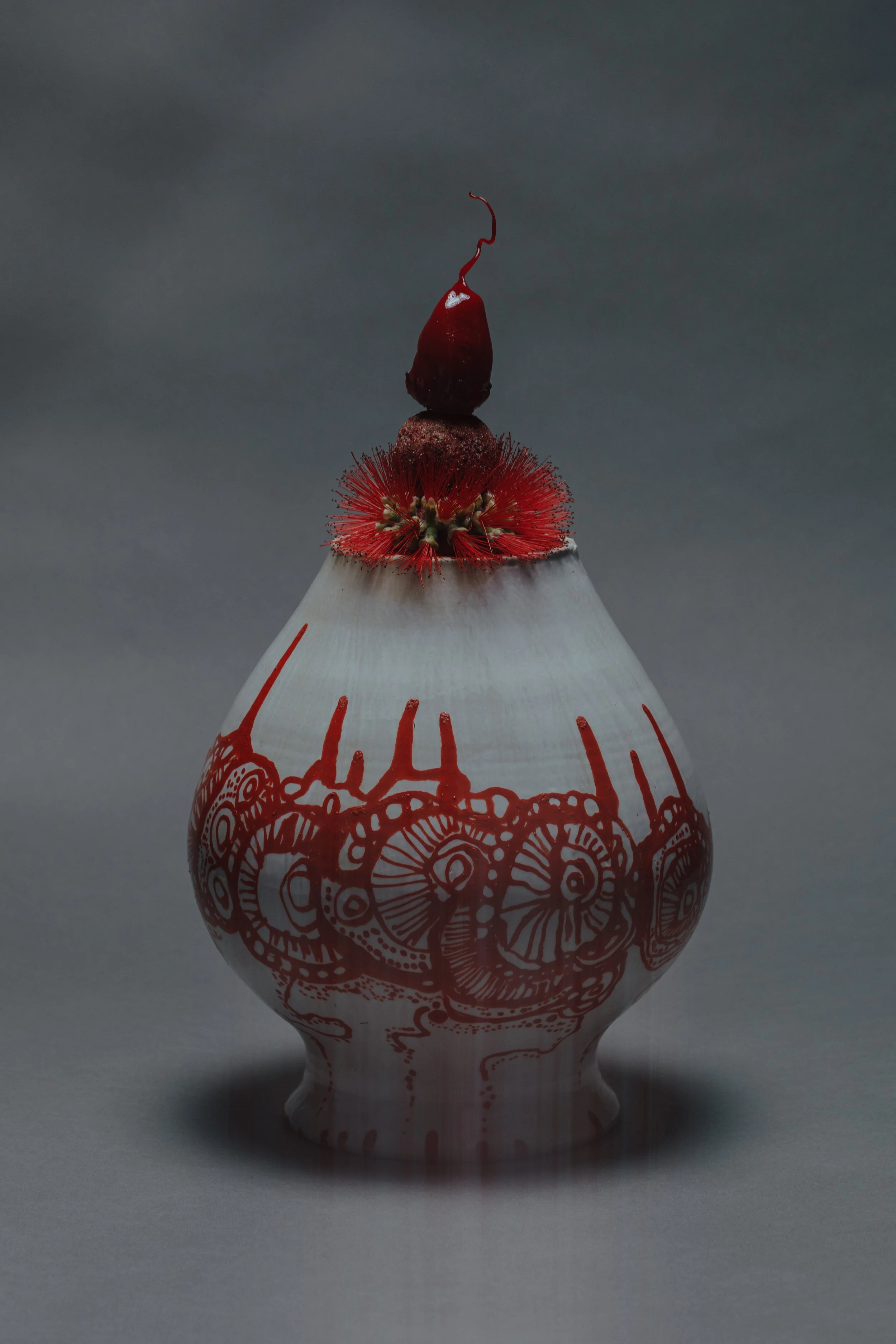 A white ceramic or porcelain vase with red floral and geometric designs. The top of the vase features a red, bead-like decoration with a dried red flower or seed pod on top. rosella, bottlebrush