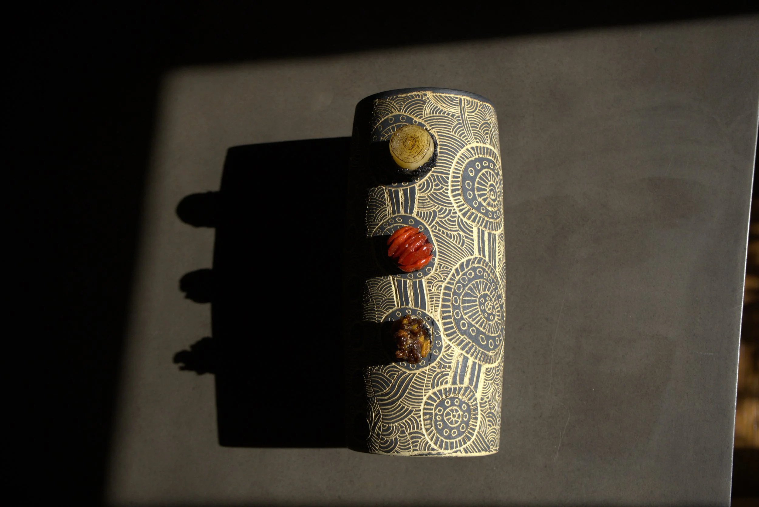 A cylindrical object with intricate gold and black geometric patterns, featuring three protruding elements in yellow, red, and black colors.