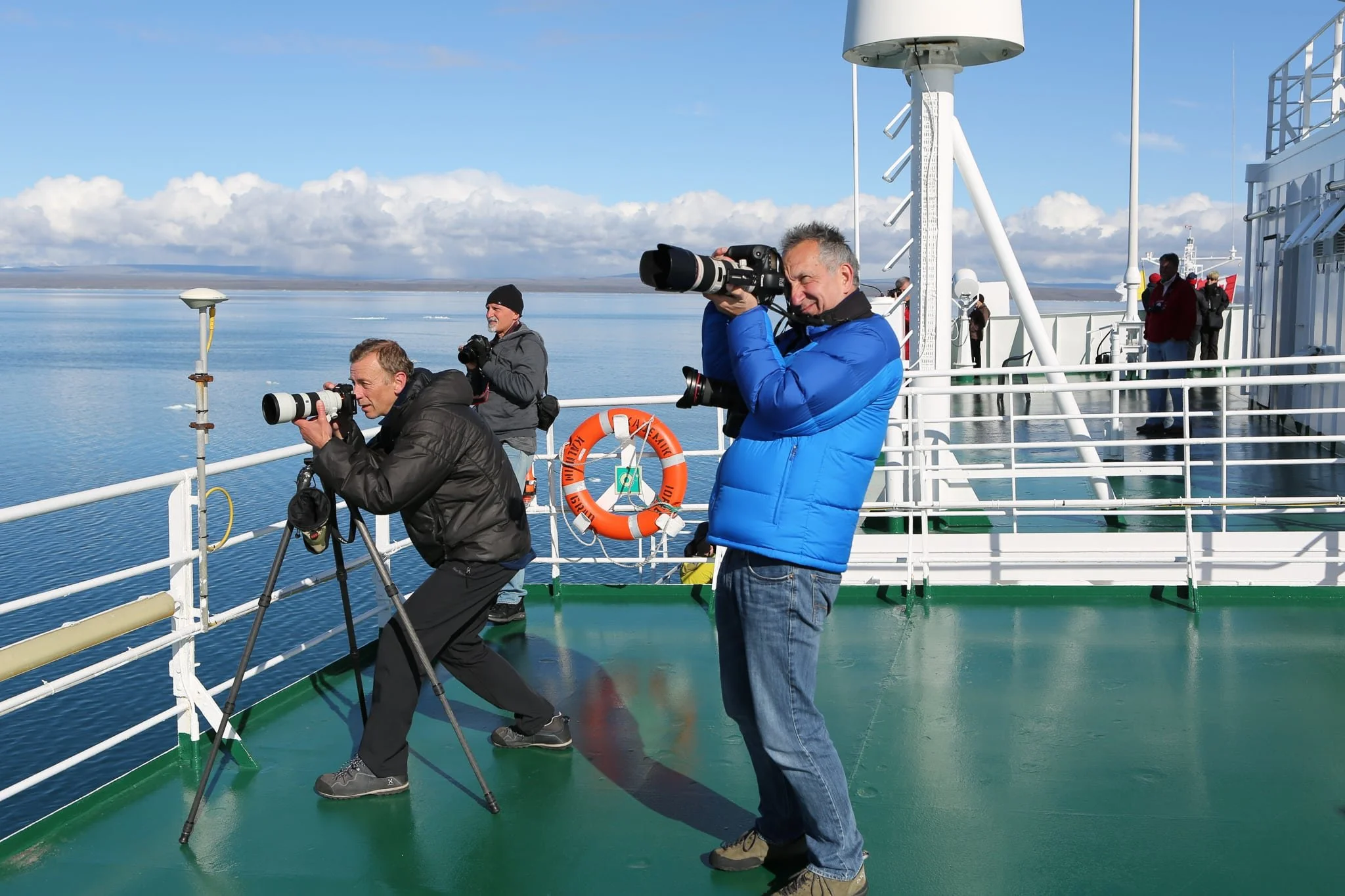 Photographers on the deck of a ship taking photos of the ocean, with a distant coastline and partly cloudy sky in the background.