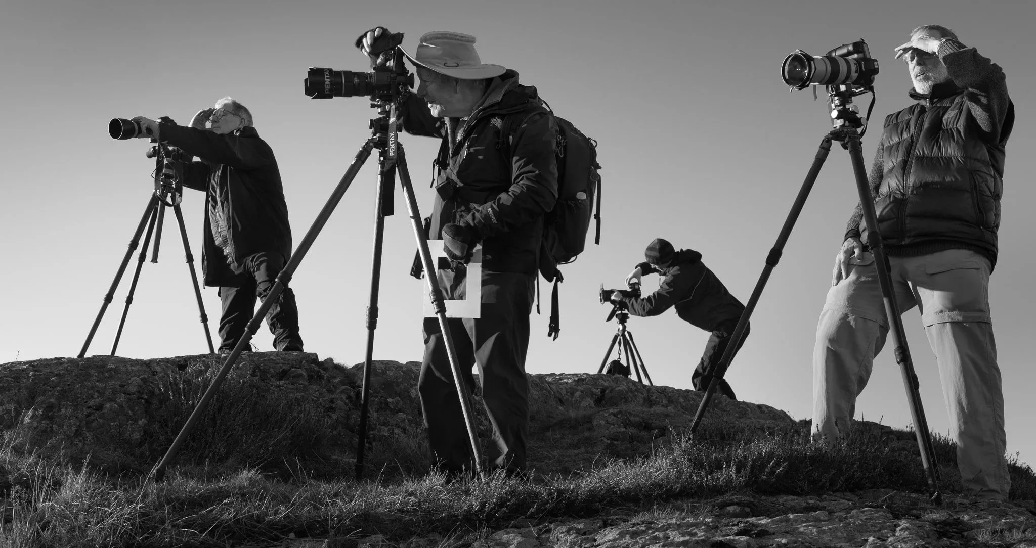 Group of four photographers on a rocky landscape, using tripods and cameras to capture a scene, with clear sky and sunlight.