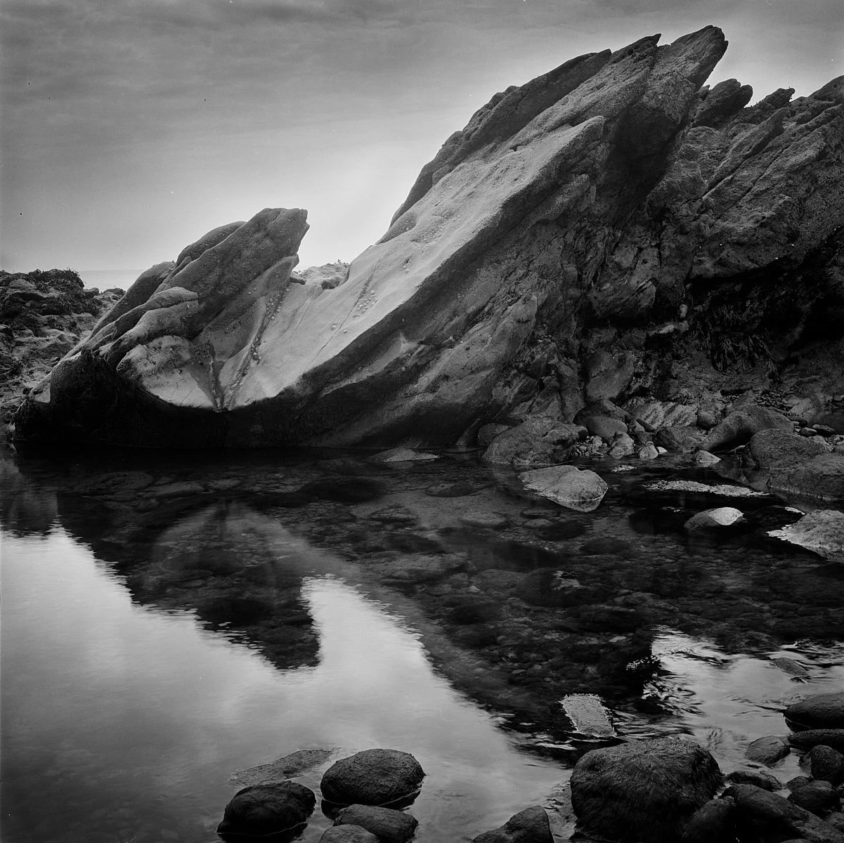 Black and white photograph of large rock formation by a calm body of water with rocky shore, showing reflections of rocks and sky.
