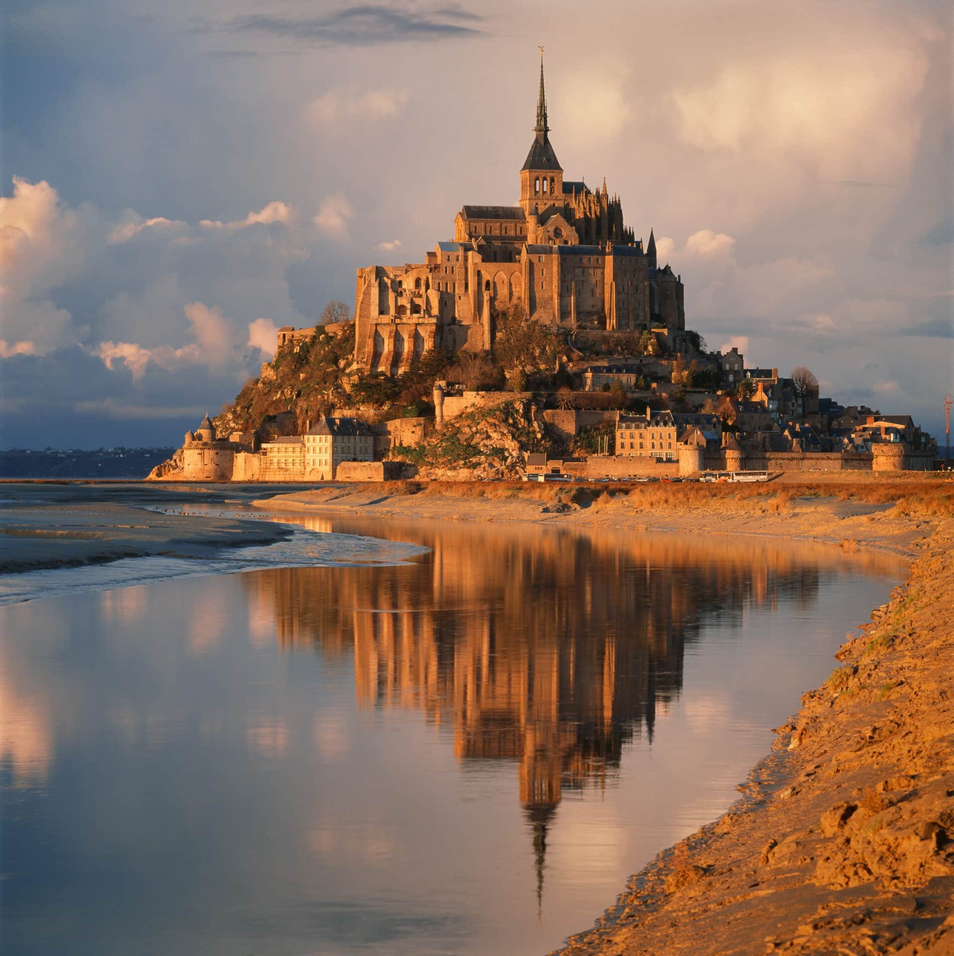 A historic church with a tall spire on a small island, surrounded by water and illuminated by warm sunlight during sunset, with clouds in the sky and scattered trees and buildings at the base of the island.