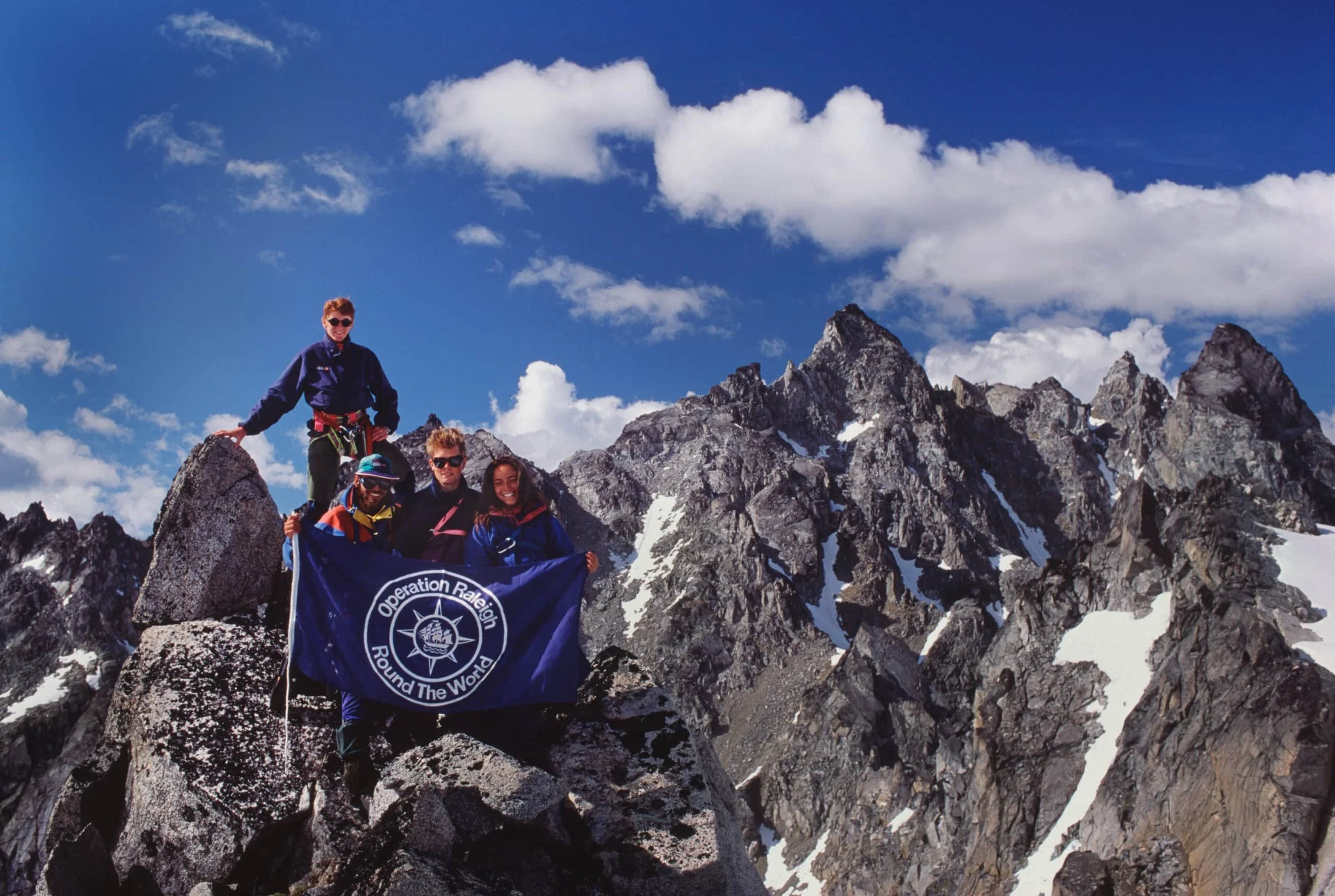 Group of five hikers on a rocky mountain peak holding a flag with the logo 'Operation Raleigh Round The World,' with snow-capped mountains in the background and a partly cloudy sky.