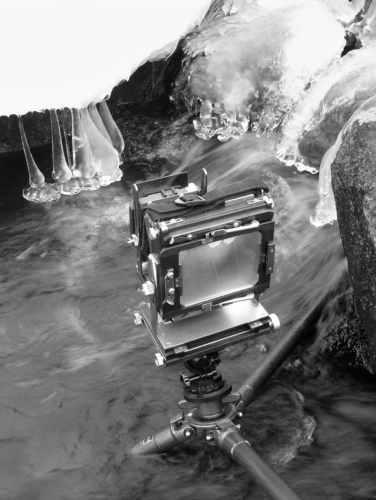 A camera mounted on a tripod in a frozen river with ice and icicles on rocks, capturing a scene of flowing water.