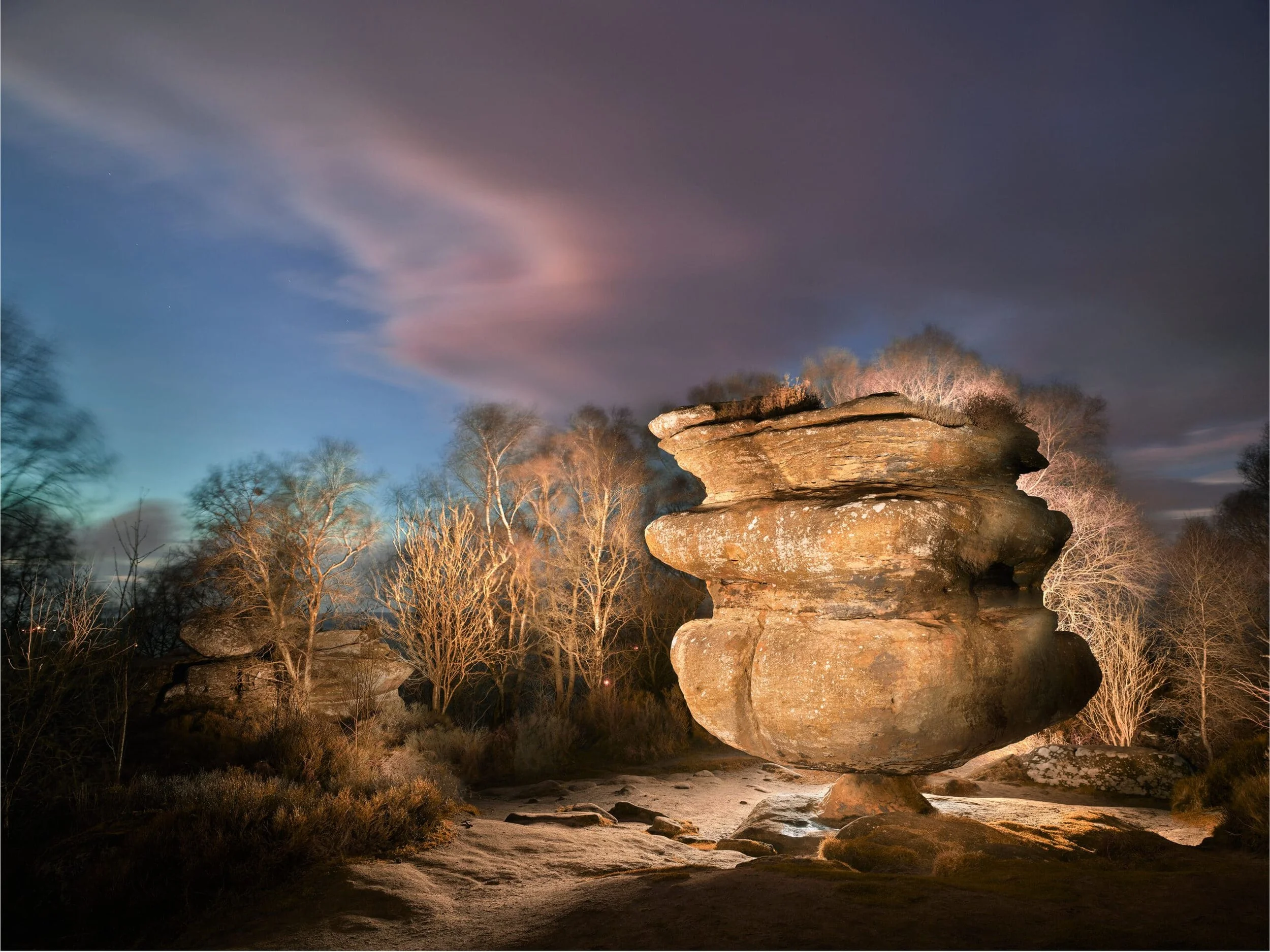 Nighttime landscape featuring large rock formation illuminated by artificial lighting, with bare trees and a softly lit trail in the foreground under a cloudy sky with hints of pink and blue.