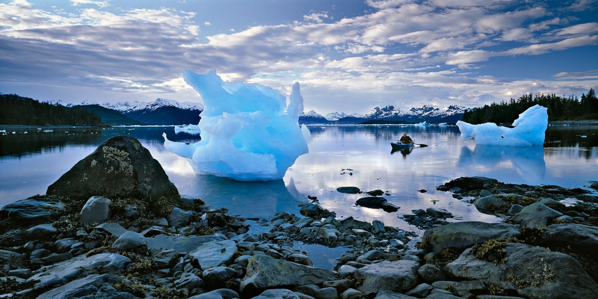 A person kayaking in a body of water with floating icebergs, snow-capped mountains, and a partly cloudy sky in the background.