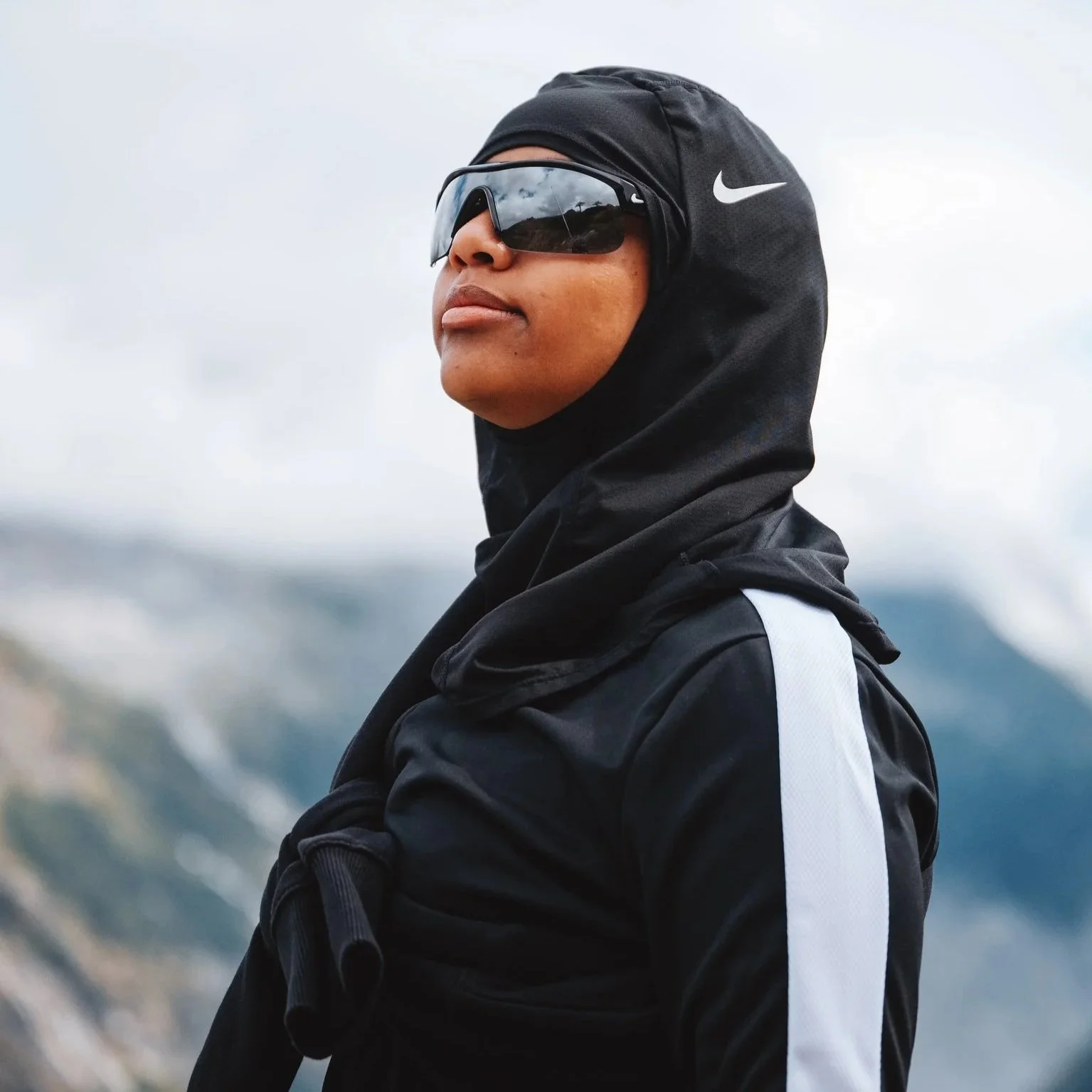 Person wearing black Nike jacket, black hood, sunglasses, and a white backpack strap outside on a cloudy day.