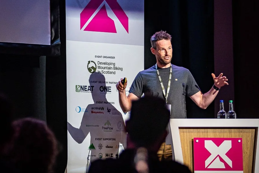A man giving a presentation at an event about mountain biking in Scotland, standing behind a podium with bottles of water, with a banner displaying event sponsors and partners