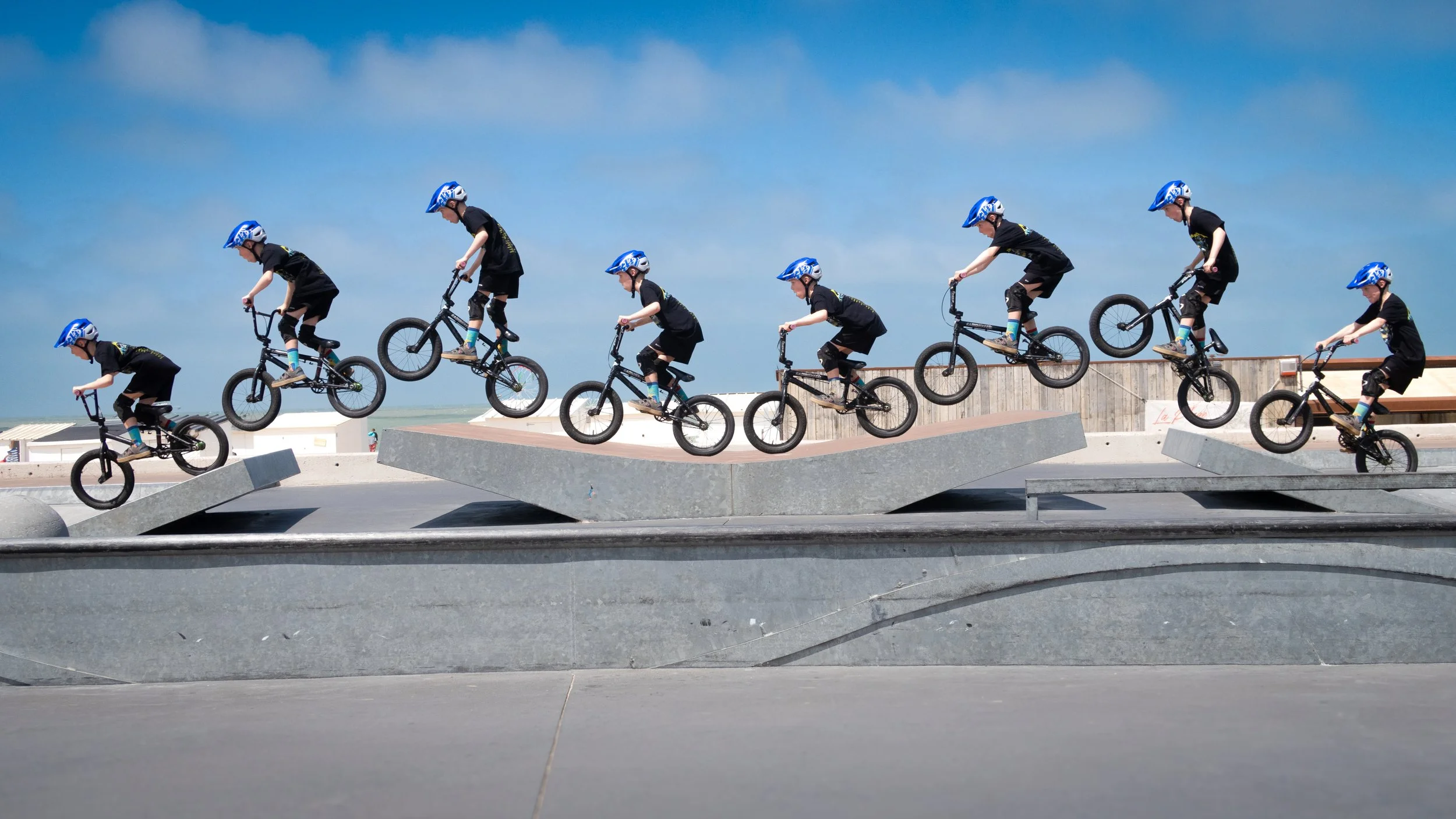 A sequence of children performing BMX bike tricks at a skate park, wearing black shirts, shorts, and blue helmets.