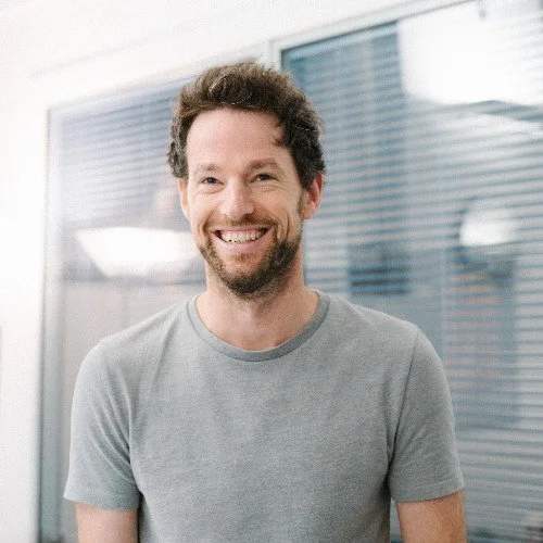 A smiling man with curly hair and beard, wearing a gray T-shirt, standing in front of glass windows.