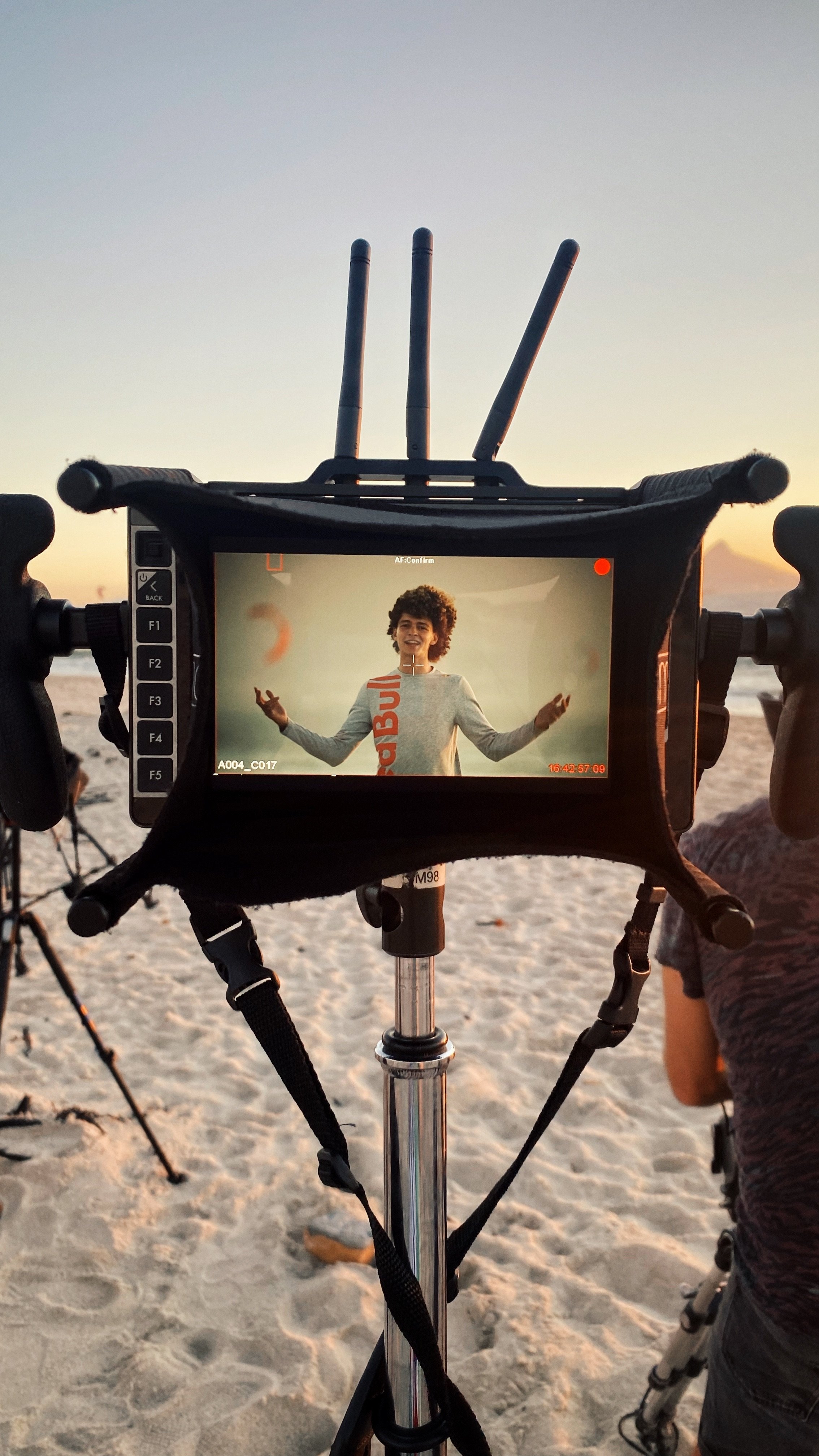 Camera viewfinder showing a person with curly hair smiling and gesturing, during sunset on a sandy beach.