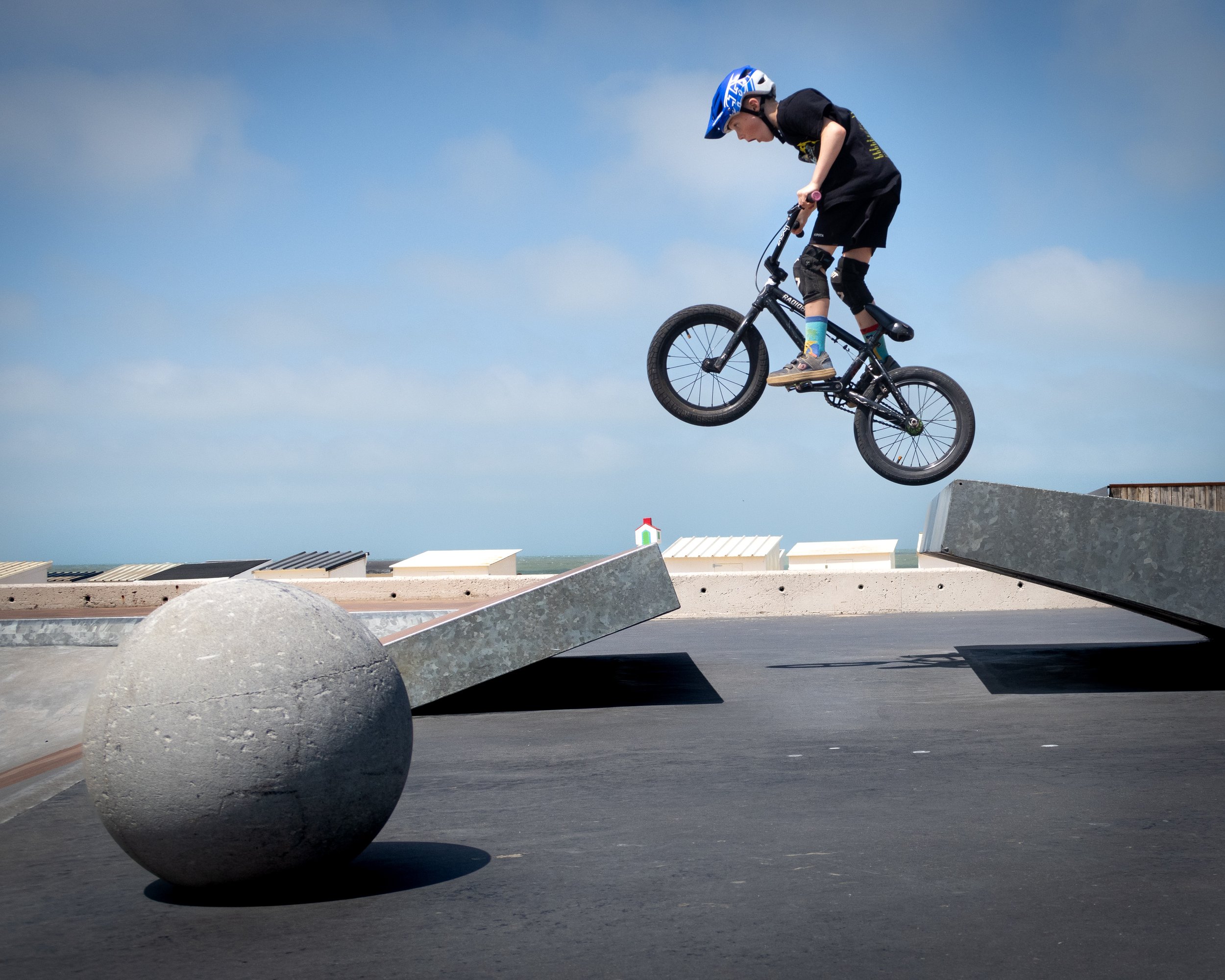Young boy wearing a helmet and knee pads riding a BMX bike up a ramp at a skate park with a blue sky in the background.