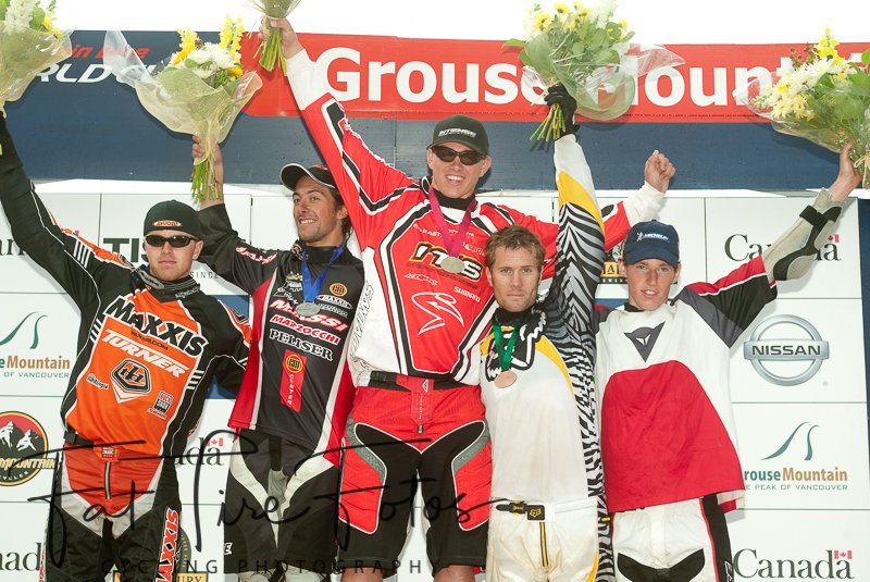 Motorsport winners' podium with five male racers holding bouquets of flowers, celebrating an event. The central figure is dressed in a red racing suit and holding a trophy, flanked by other racers in racing gear, all smiling. The background displays logos and sponsor banners.