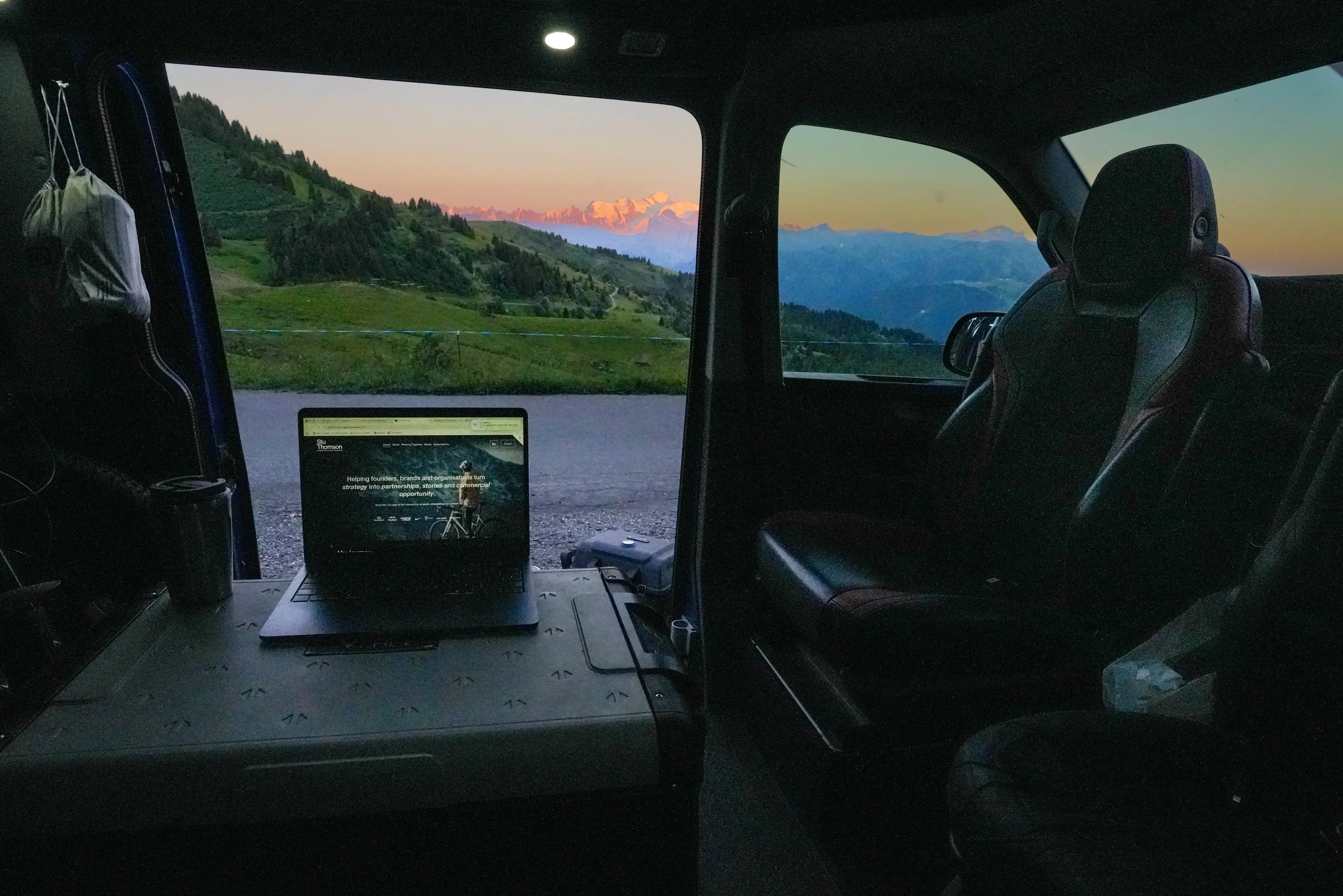 Inside a van with a view of distant mountains and a green hillside at dusk, a laptop is open on a desk, a reusable coffee cup, and other small items are on the desk.