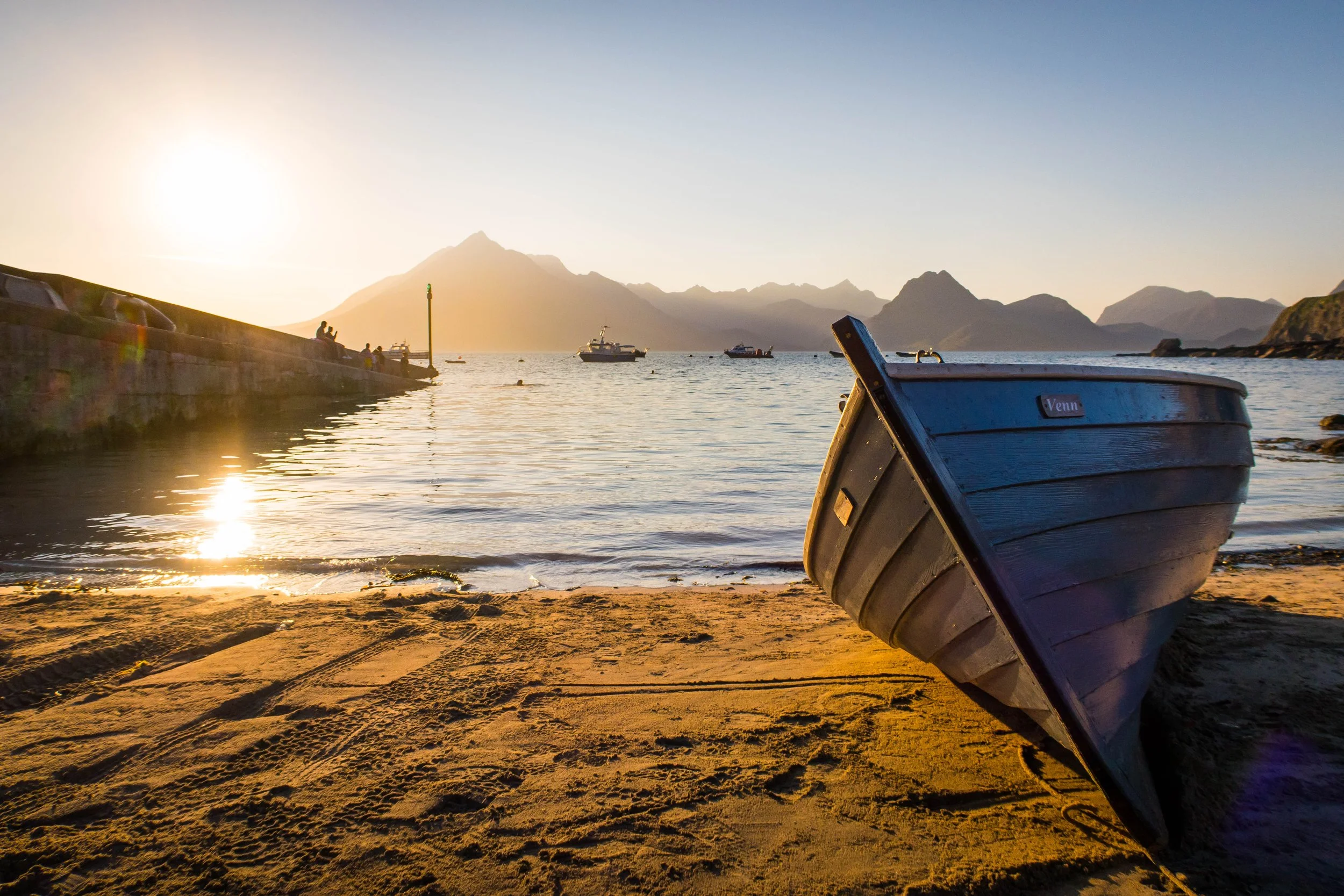 A boat on the sandy shore with mountains in the background during sunset, with a stone pier and people sitting and fishing.