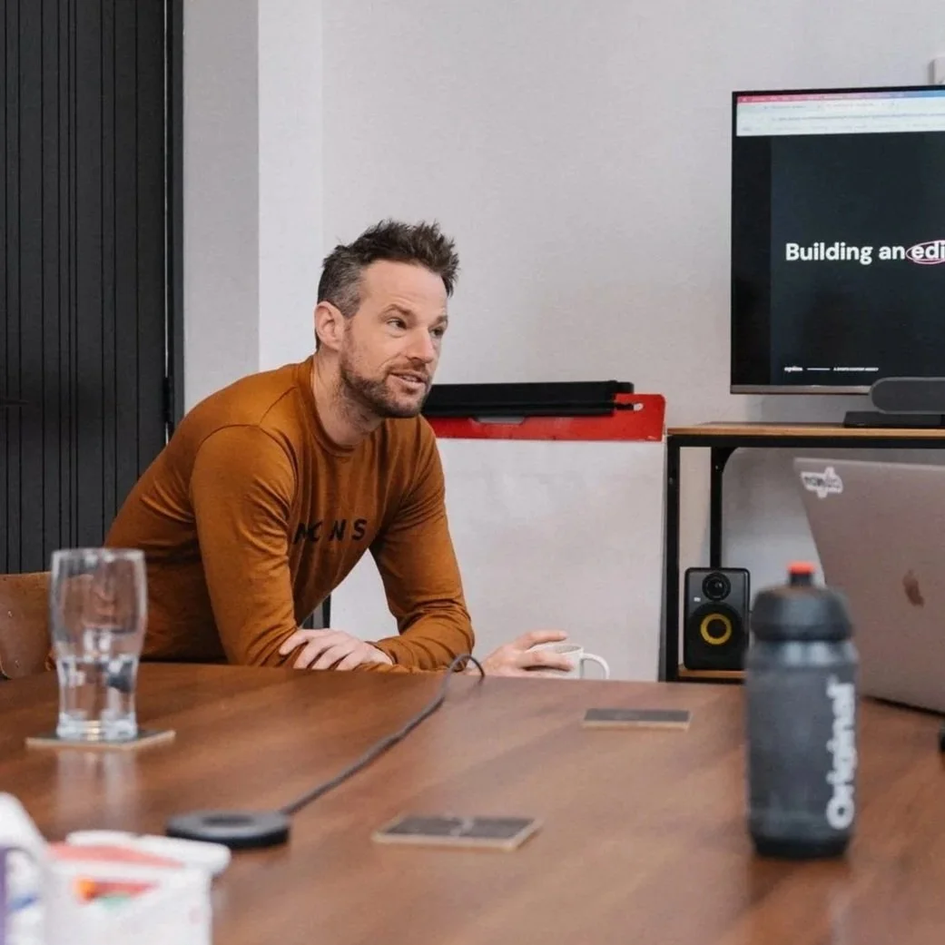 A man with short, spiky hair and a beard, wearing a brown long-sleeve shirt, sitting at a wooden conference table with a glass of water and a bottle in front of him. He is in a meeting room with a large monitor displaying a presentation, and various electronic equipment and office supplies are visible.