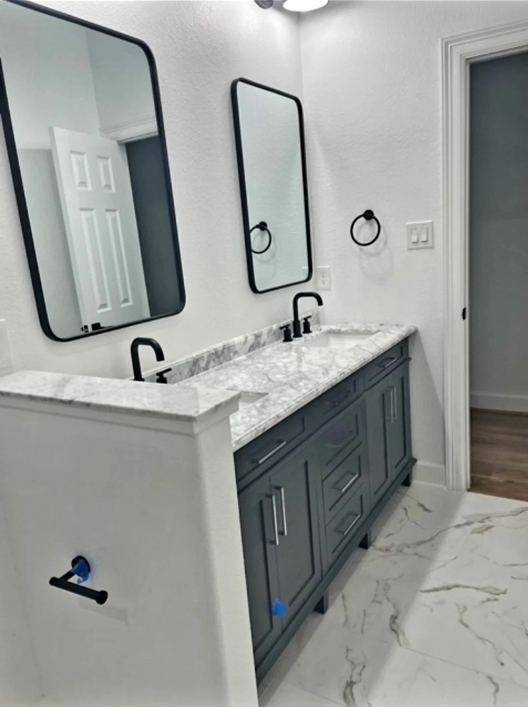 Bathroom with dual sinks, marble countertop, gray vanity, two black framed mirrors, and white walls.