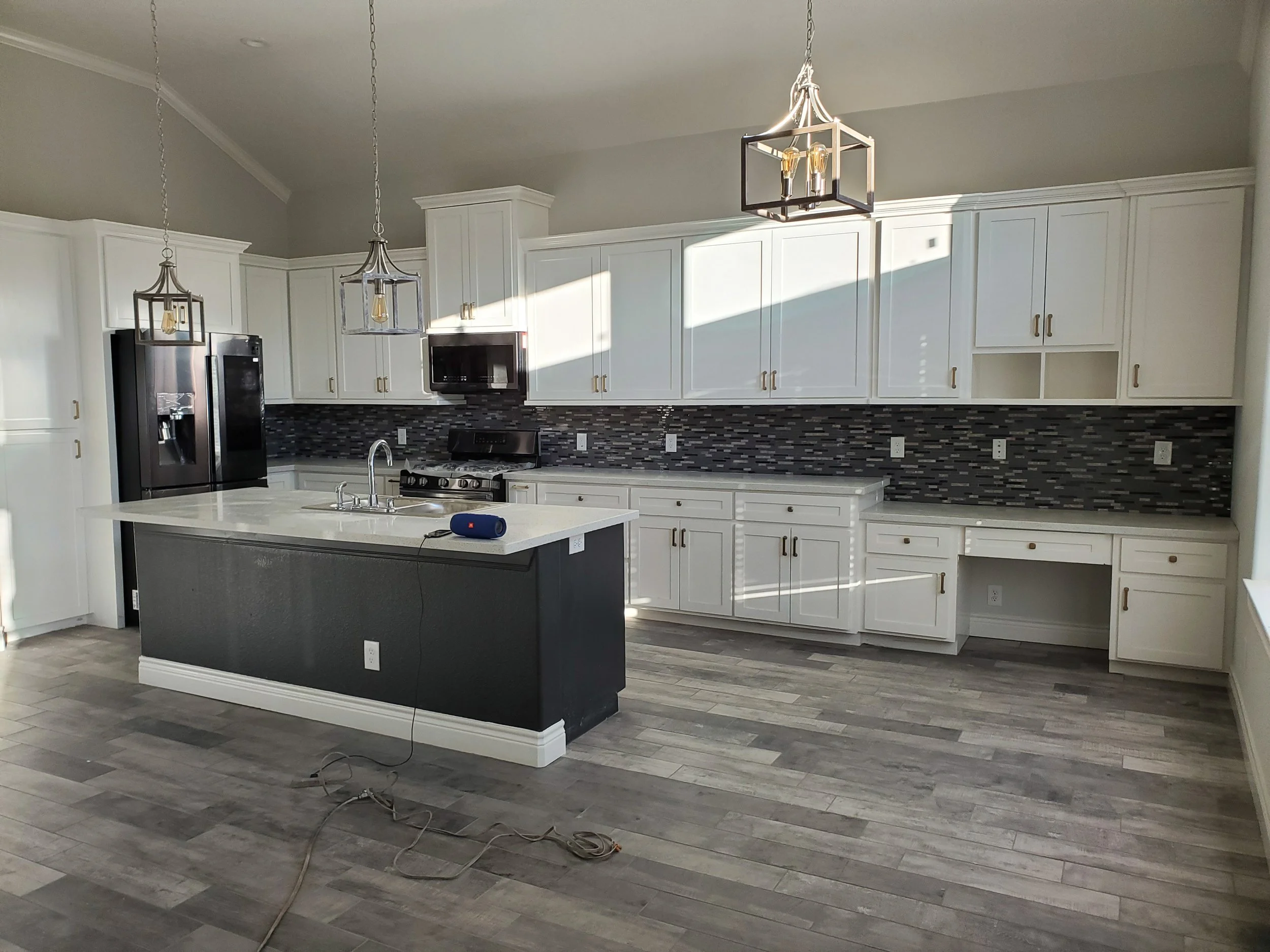 Modern kitchen with white cabinets, black backsplash, gray countertops, stainless steel appliances, and a central island with a sink. Two pendant lights hang from the ceiling.