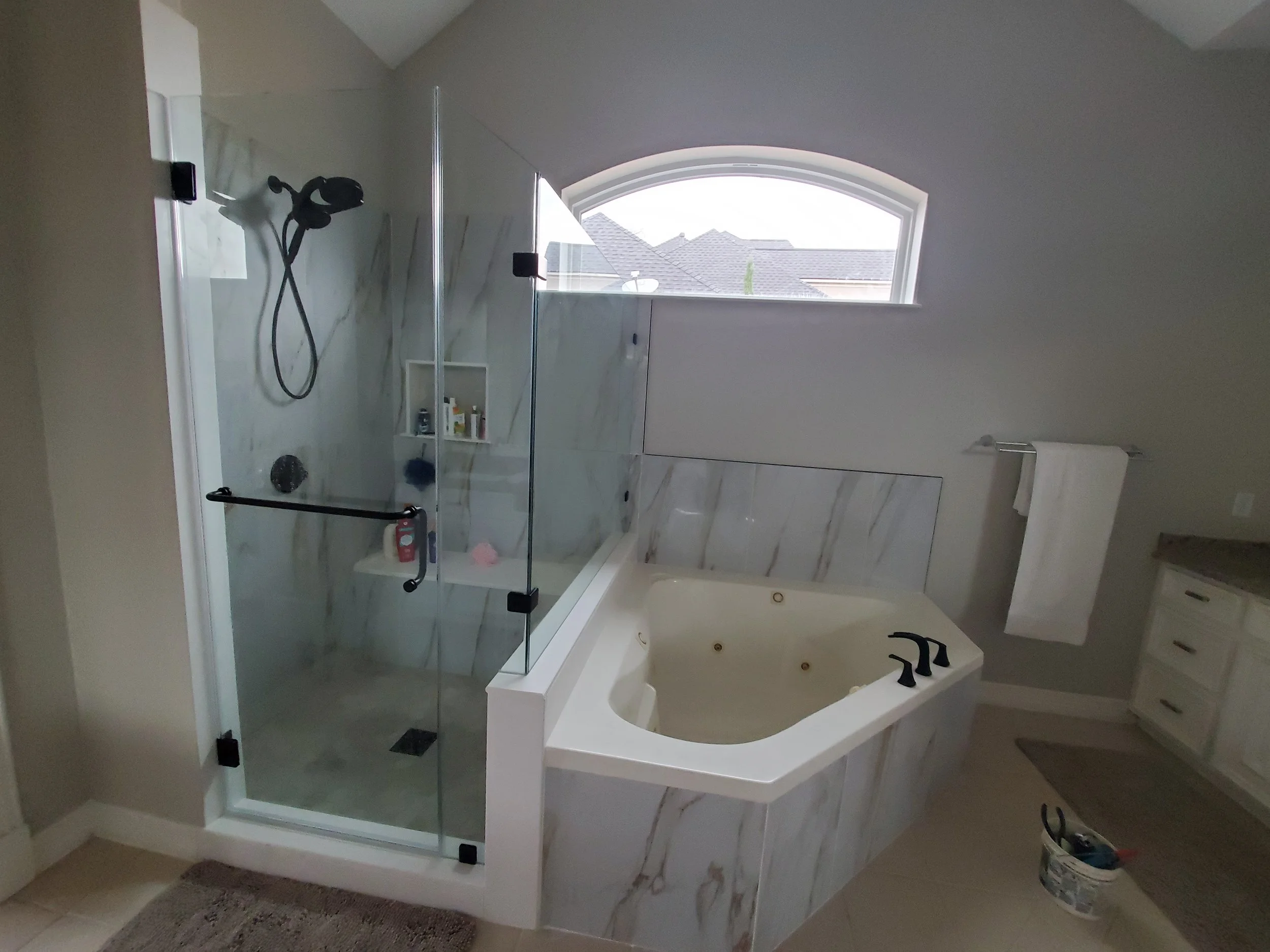 A modern bathroom with a glass-enclosed shower, a window with a view of rooftops, and a bathtub with black fixtures.
