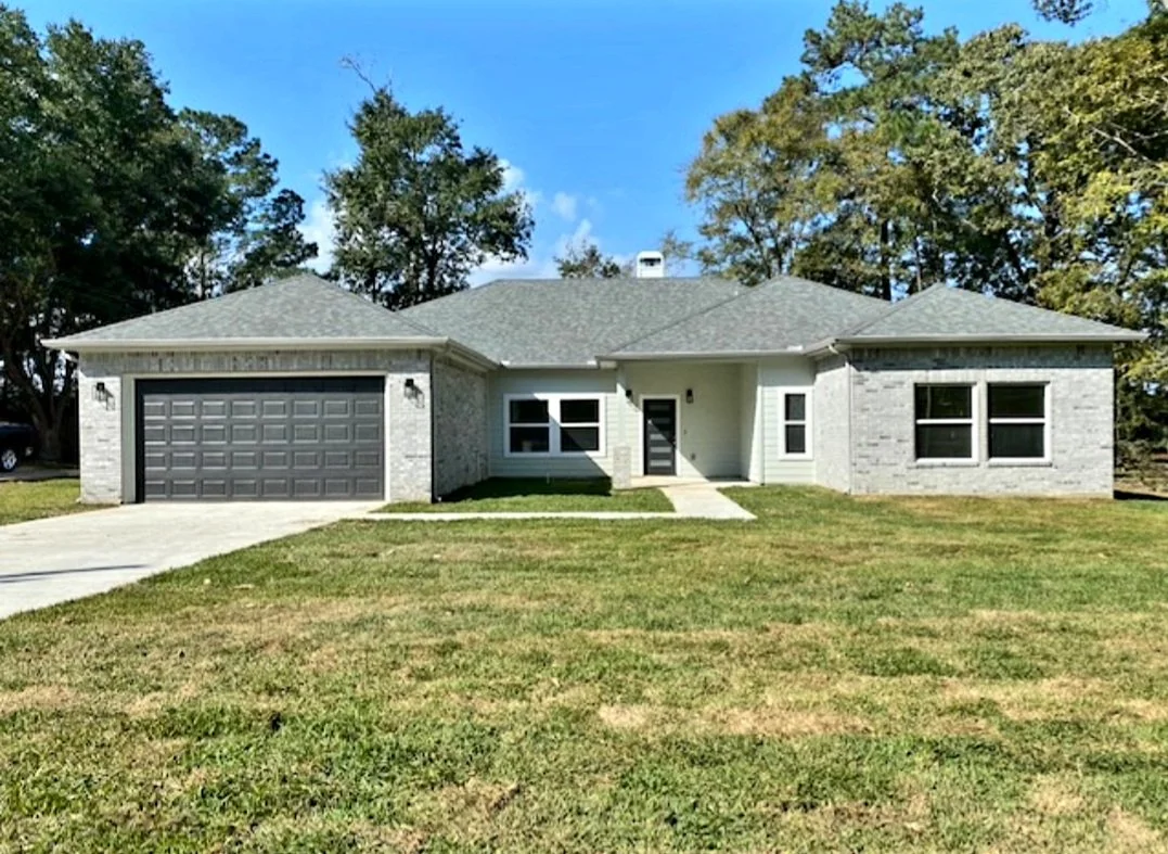 A modern single-story house with a gray exterior, a two-car garage, and a well-maintained lawn, surrounded by trees under a blue sky.