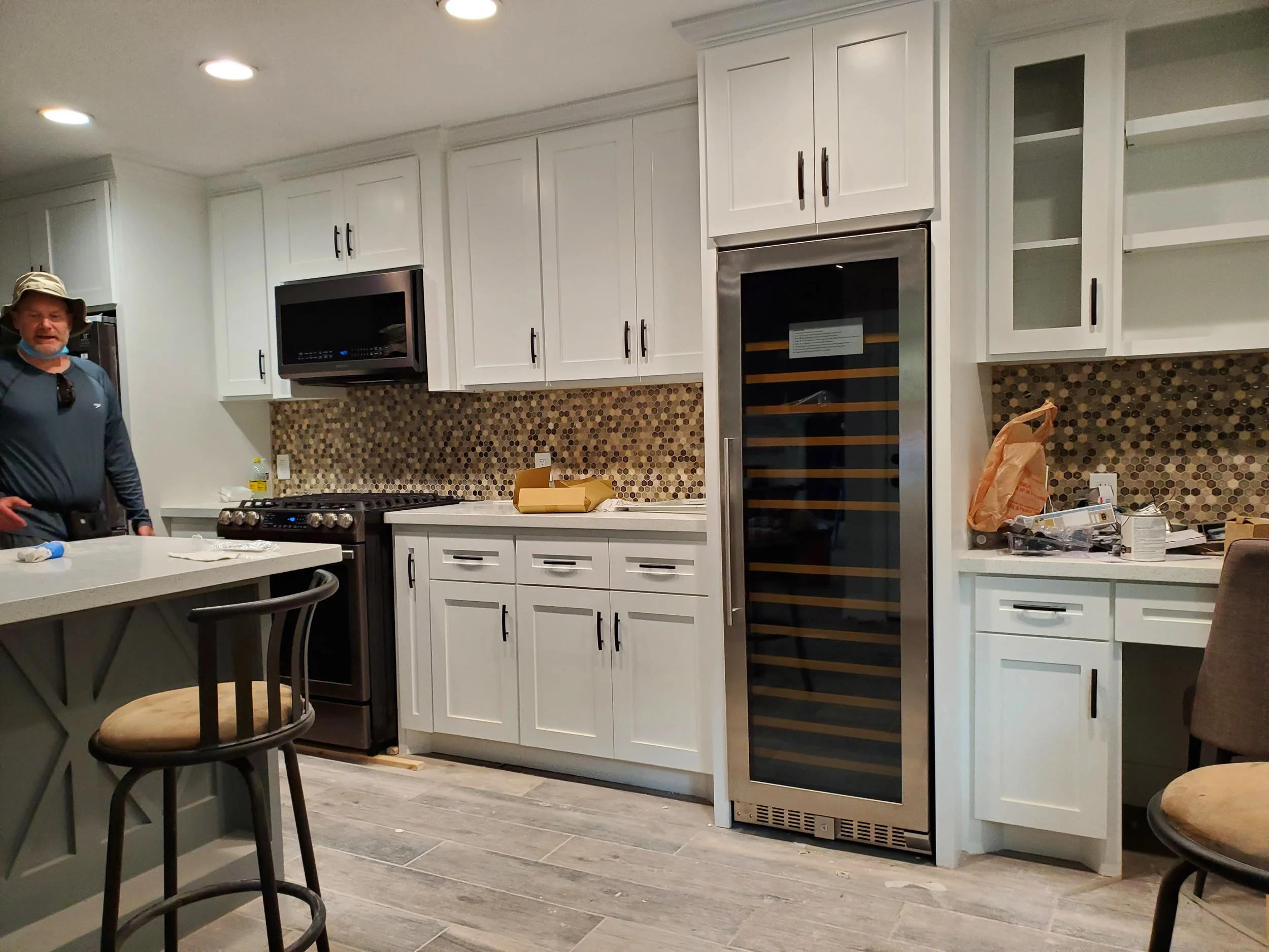 A kitchen under remodeling with white cabinets, a built-in wine fridge, a stove, and a microwave. There are chairs and various construction materials on the counters.