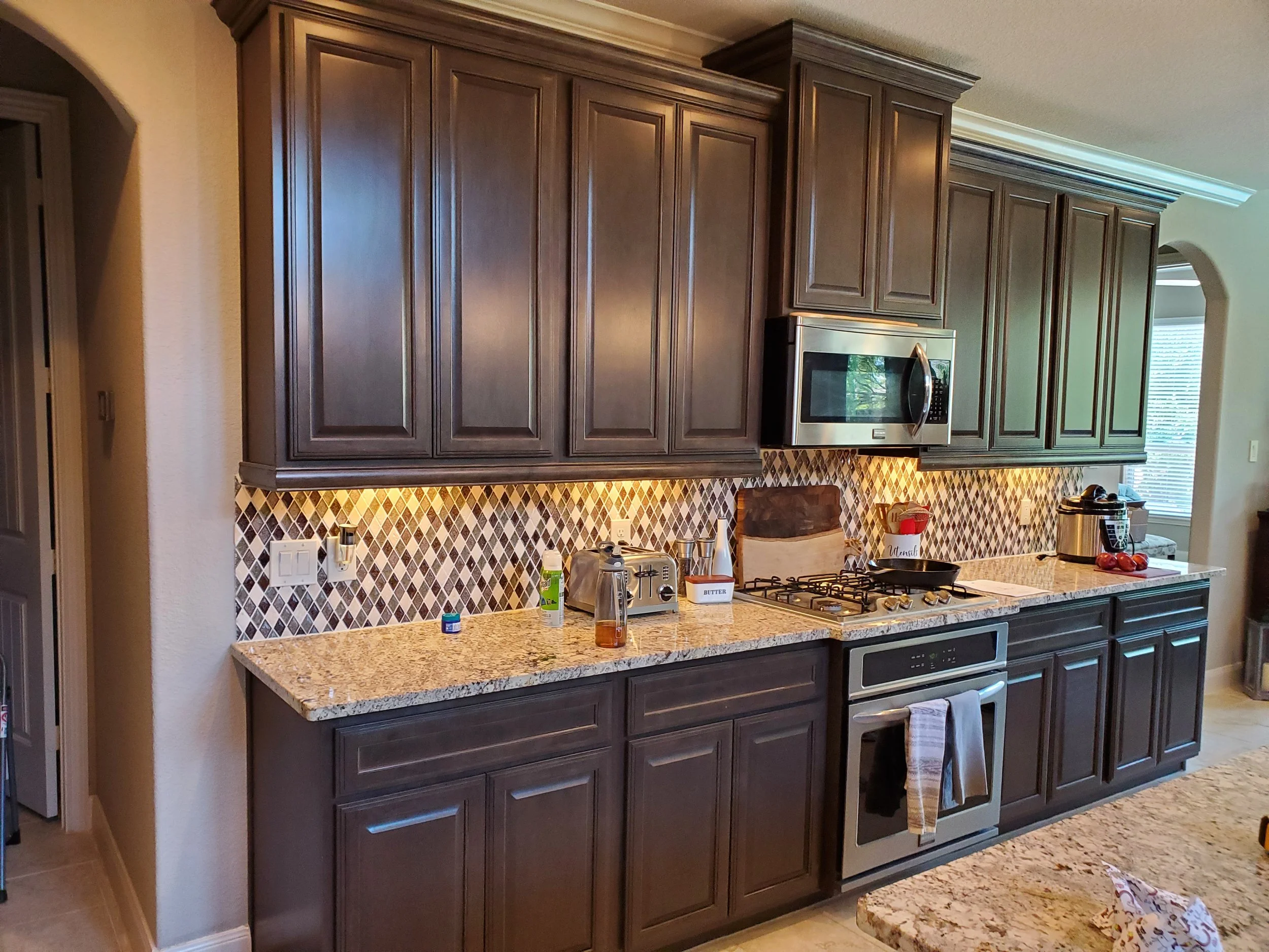 Kitchen with dark wooden cabinets, granite countertop, stainless steel microwave oven, stove, and colorful diamond-shaped backsplash tiles.