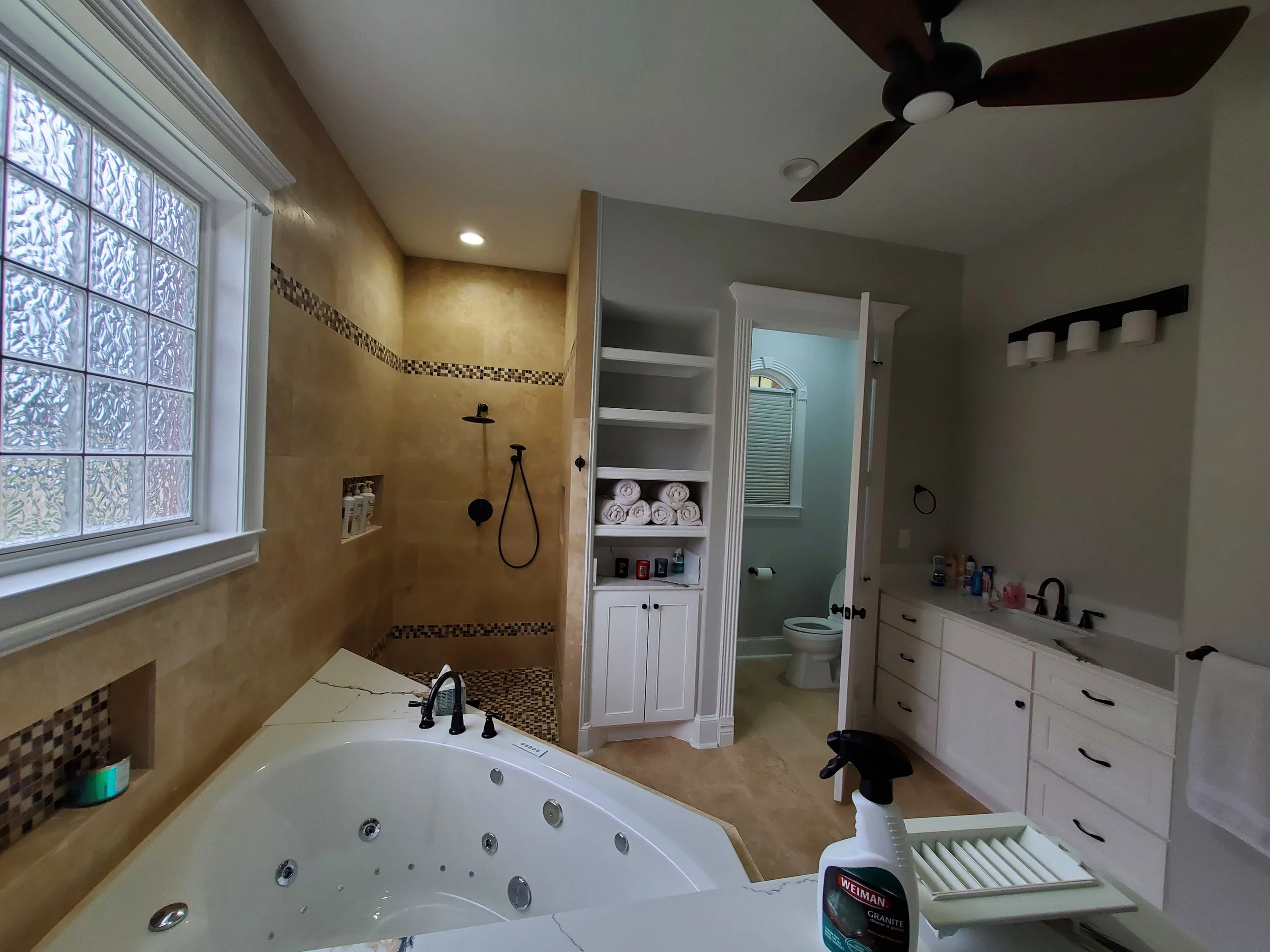 A bathroom with a large window made of glass blocks, a soaking bathtub with whirlpool jets, a shower area with a rainfall showerhead, a shelf with rolled towels, and a toilet. The room has beige tiled walls and a ceiling fan.